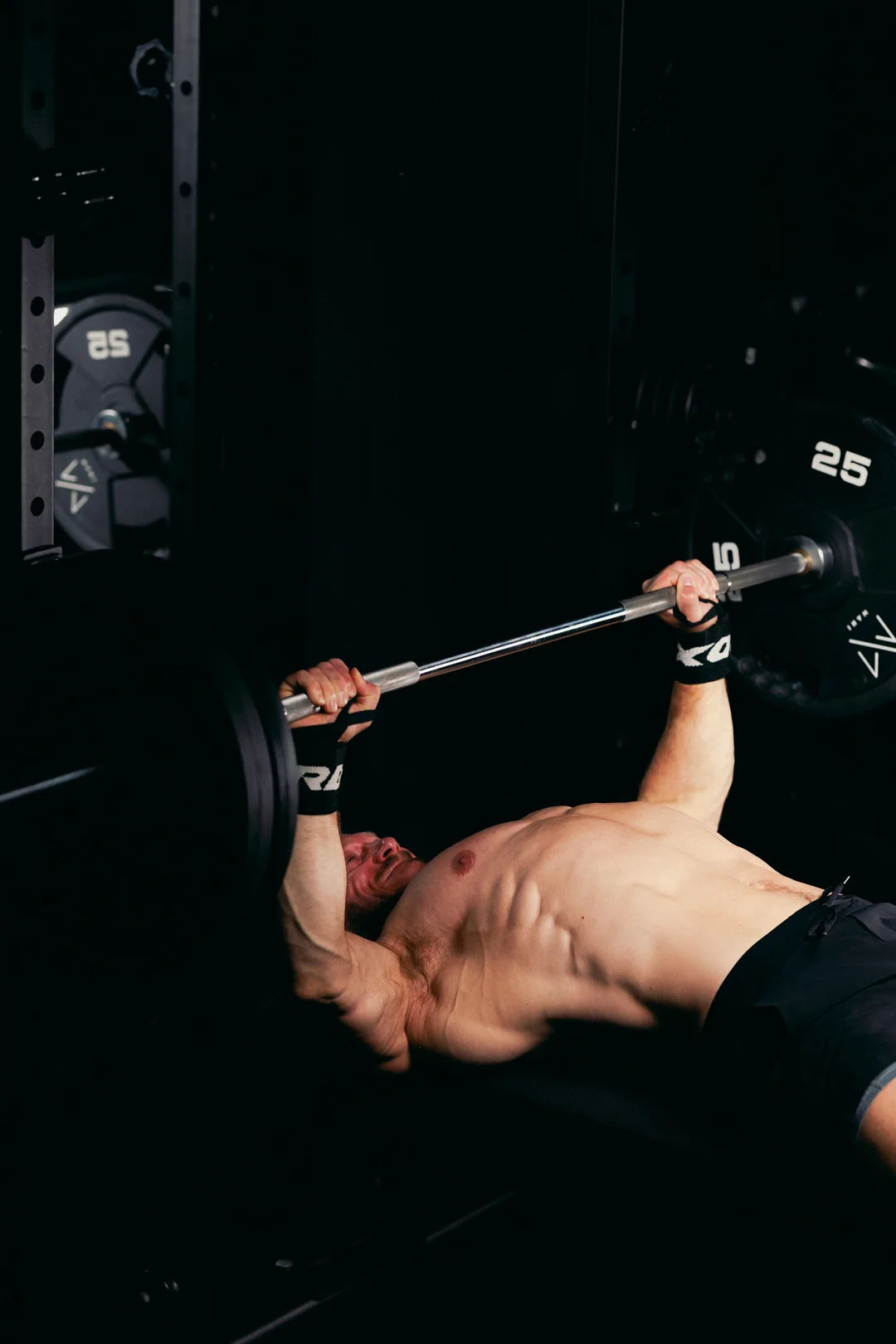 Shirtless man performing a bench press in a dark gym with dramatic lighting.