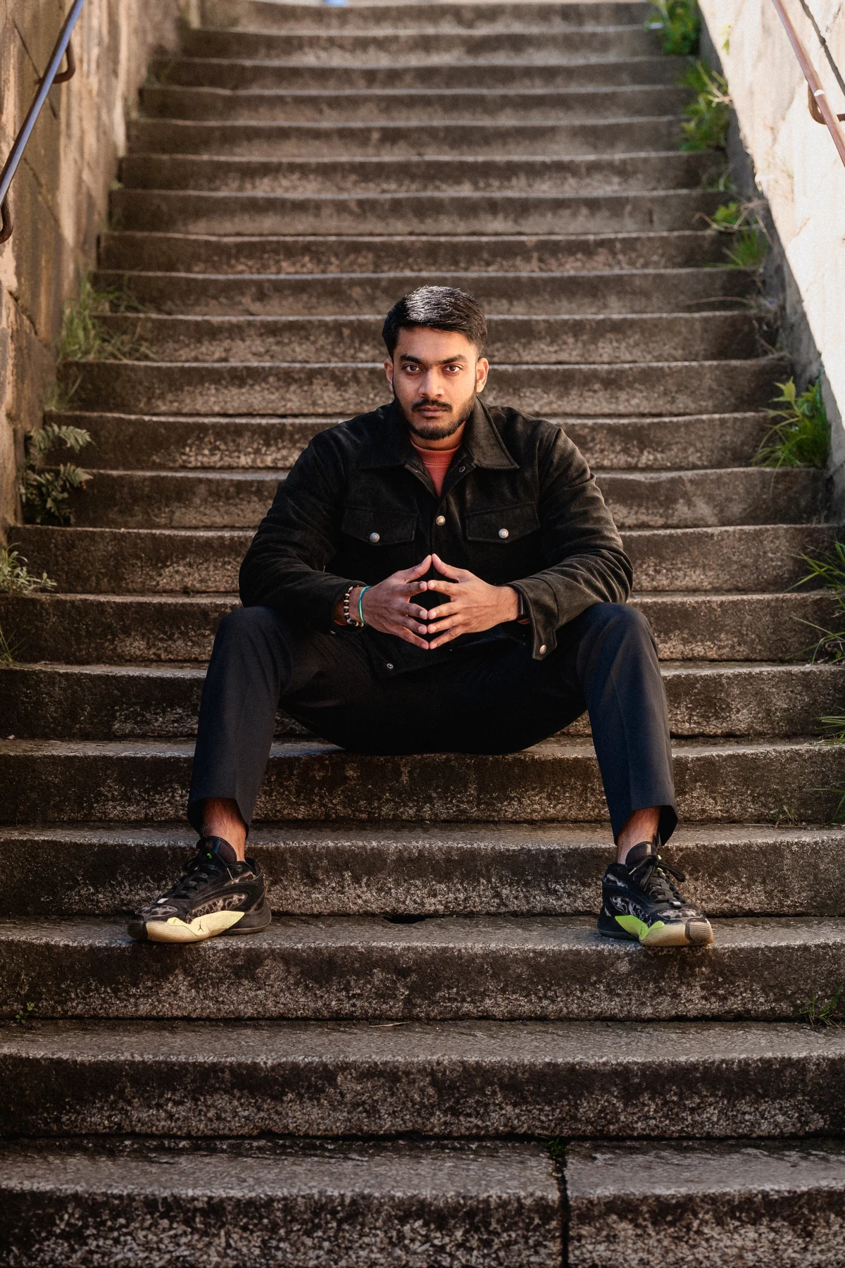 A man in a black jacket and trousers sitting on stone stairs, looking directly at the camera with a serious expression.