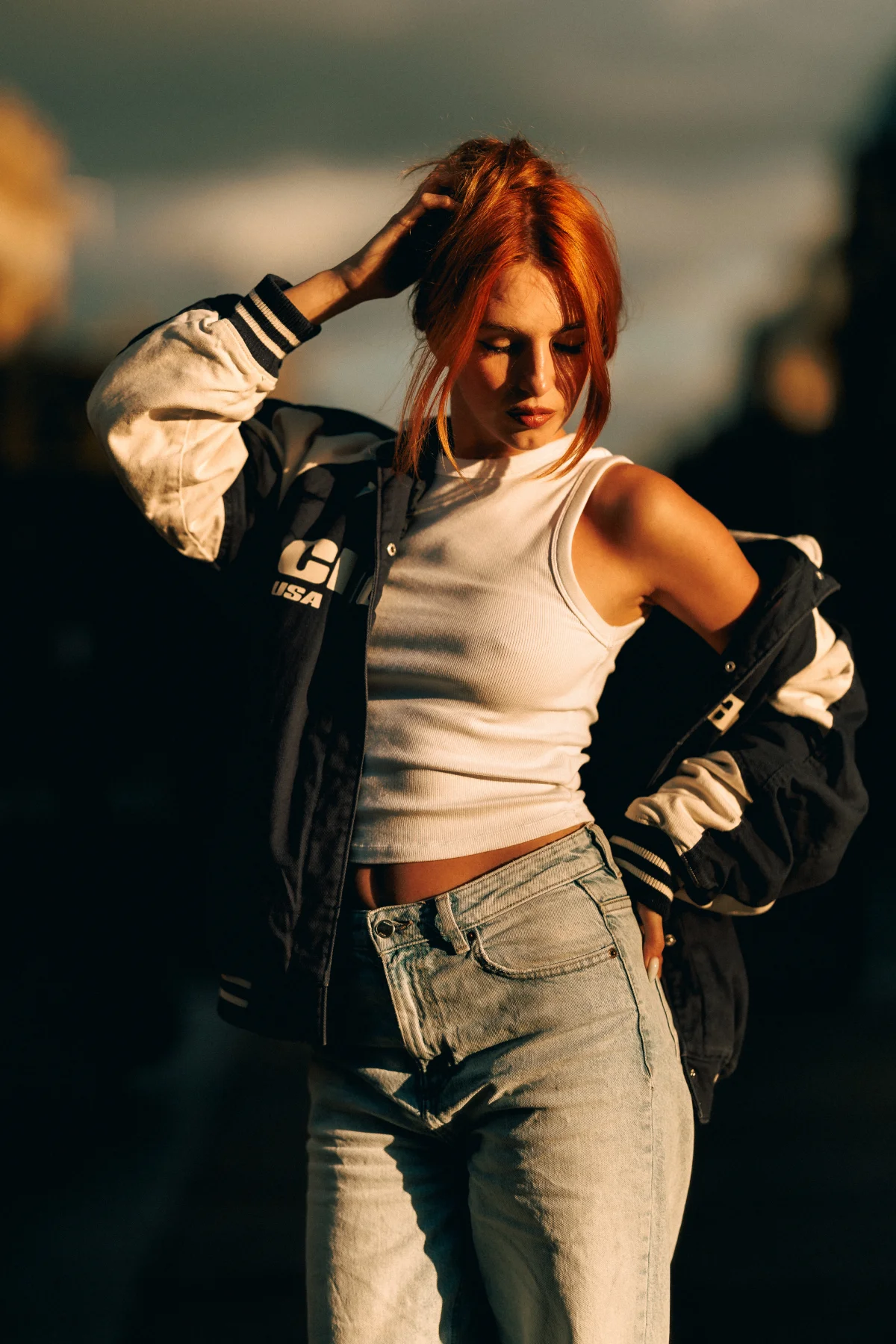 Woman with red hair in a varsity jacket and white crop top posing in golden hour light.