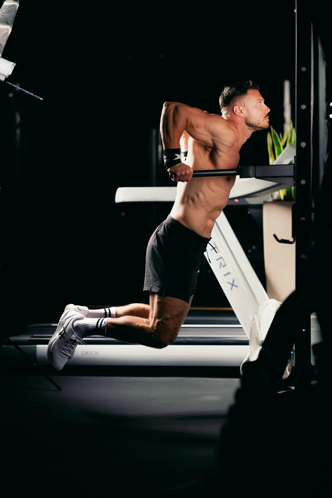 A muscular shirtless man performing a dip exercise on gym equipment with dramatic studio lighting.