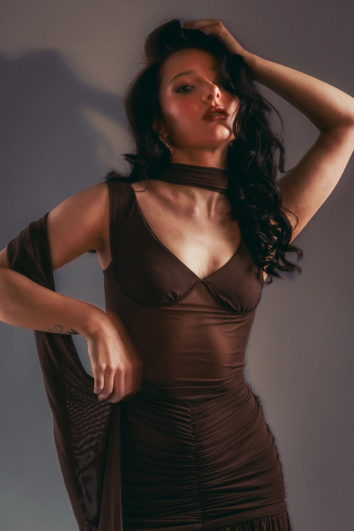 Woman with long dark wavy hair in a brown form-fitting dress posing against a grey studio background.