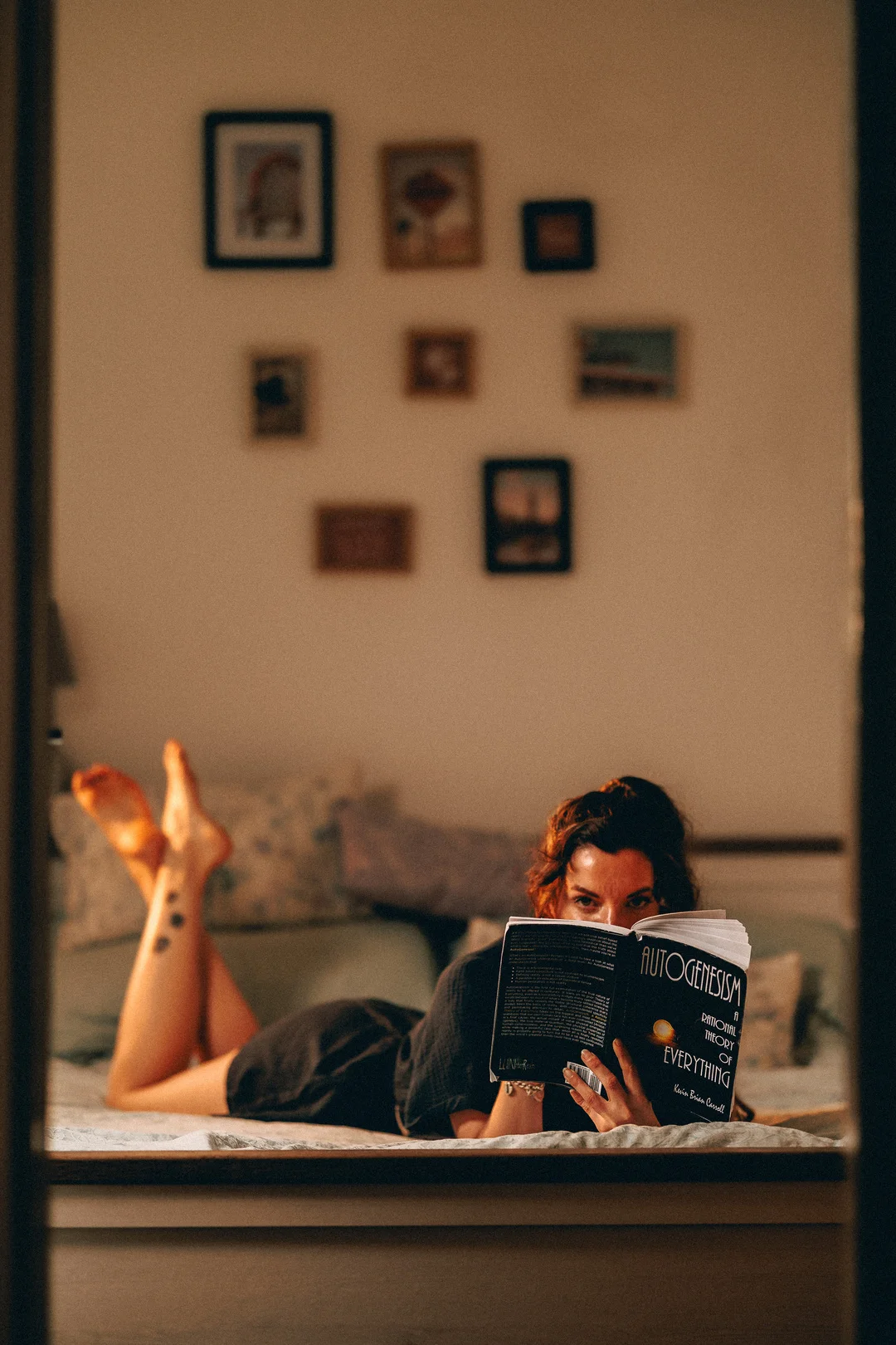 A woman lying on a bed reading a book, viewed through a doorway in a cozy, dimly lit room.