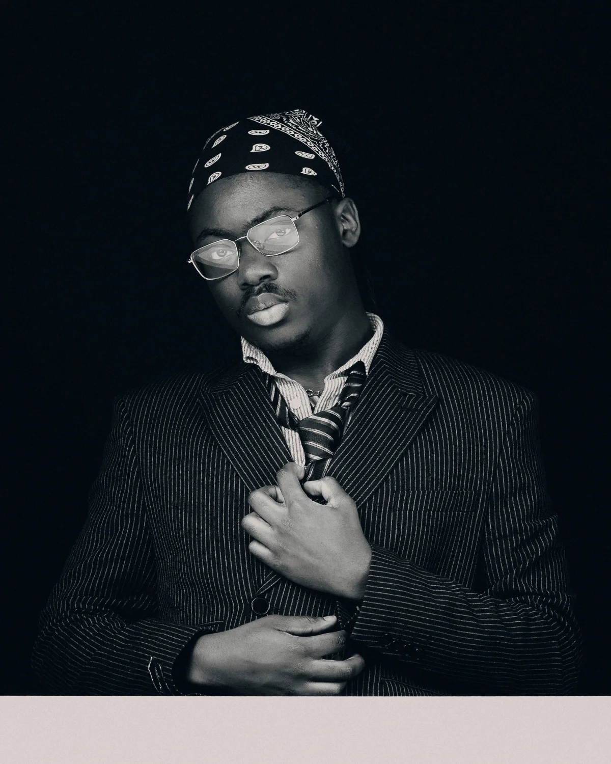 Black and white studio portrait of a man in a pinstripe suit, glasses, and a bandana.