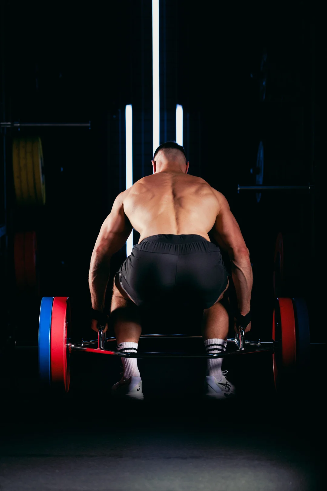 Rear view of a muscular man preparing to lift a barbell in a dark gym with dramatic vertical lighting.