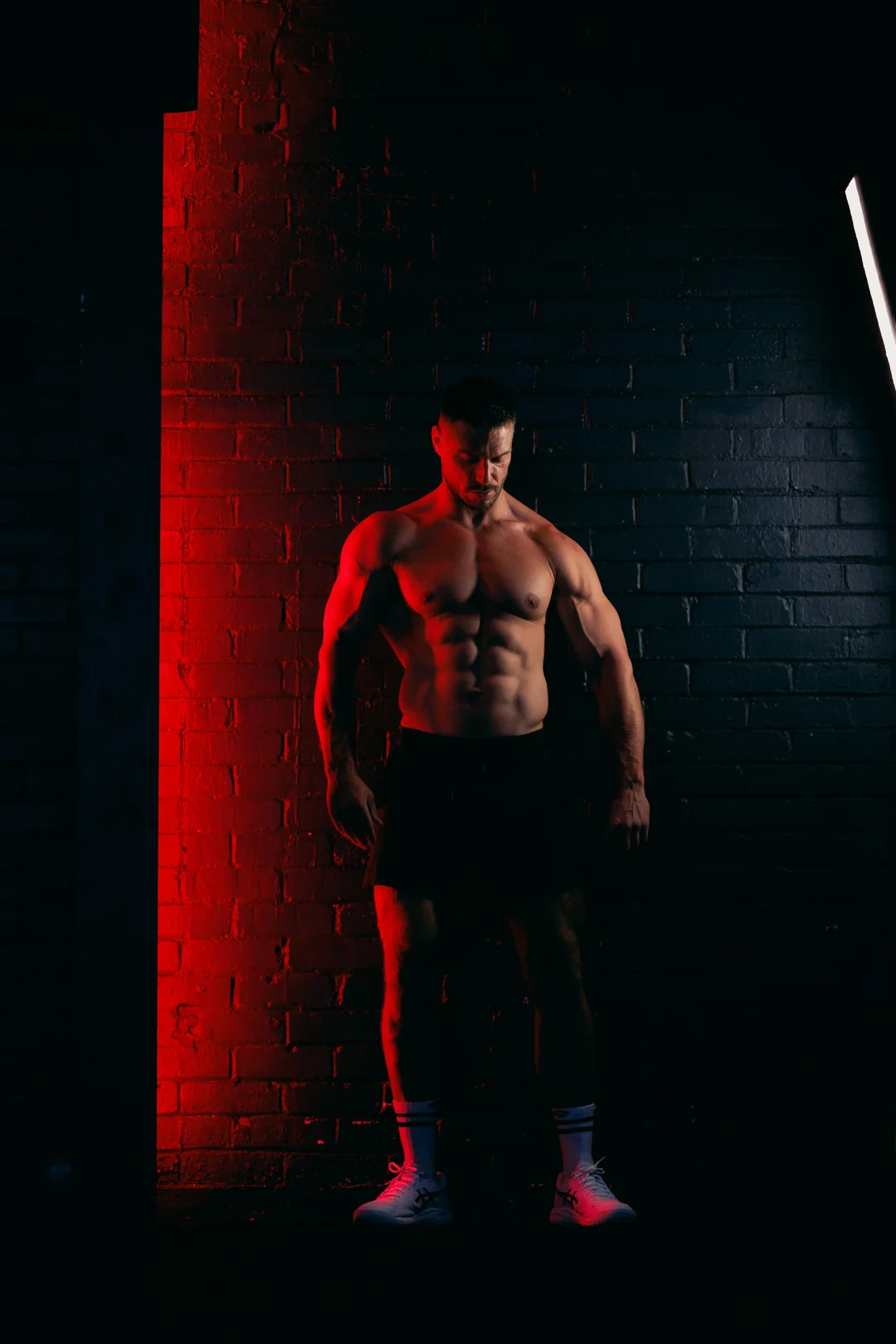 Muscular shirtless man posing against a dark brick wall with dramatic red and white lighting.