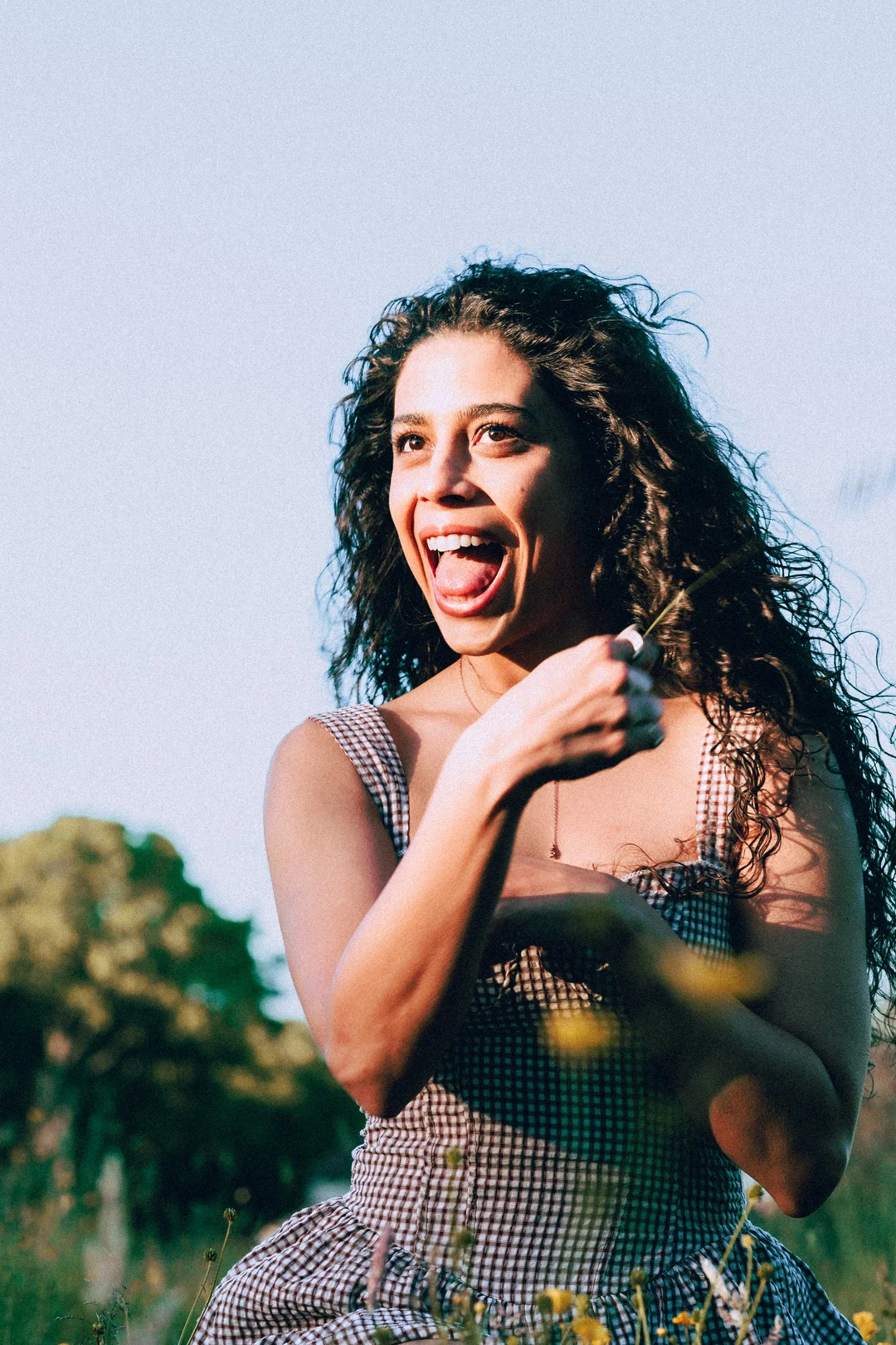 A joyful woman with curly hair in a checkered dress laughing in a sunny outdoor field.