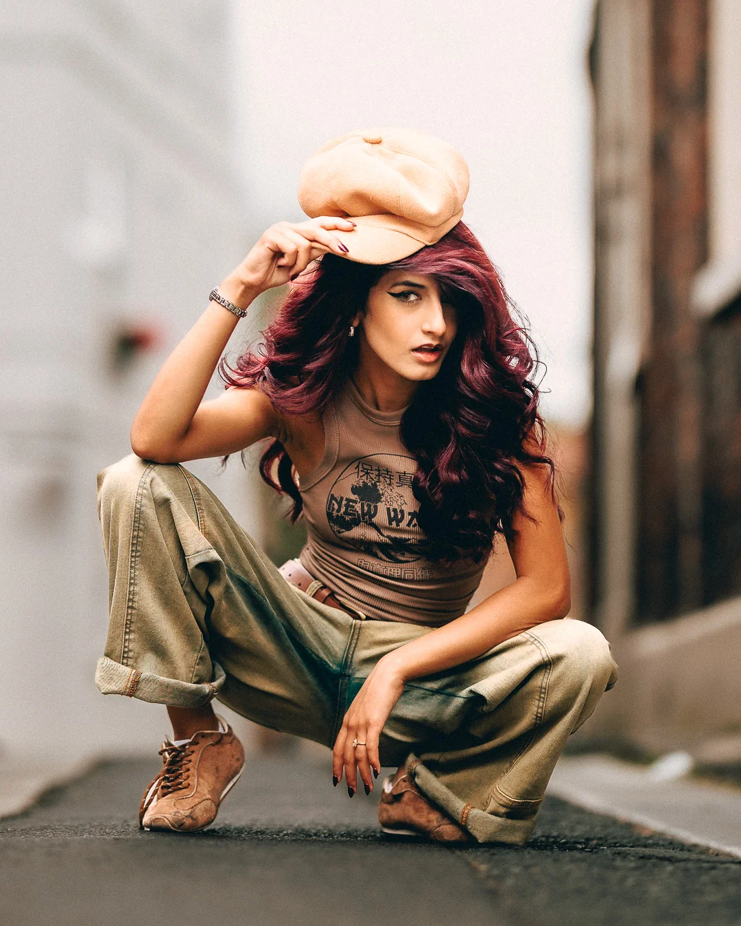A woman with long red hair in a tan cap and urban attire crouching in a city alley.