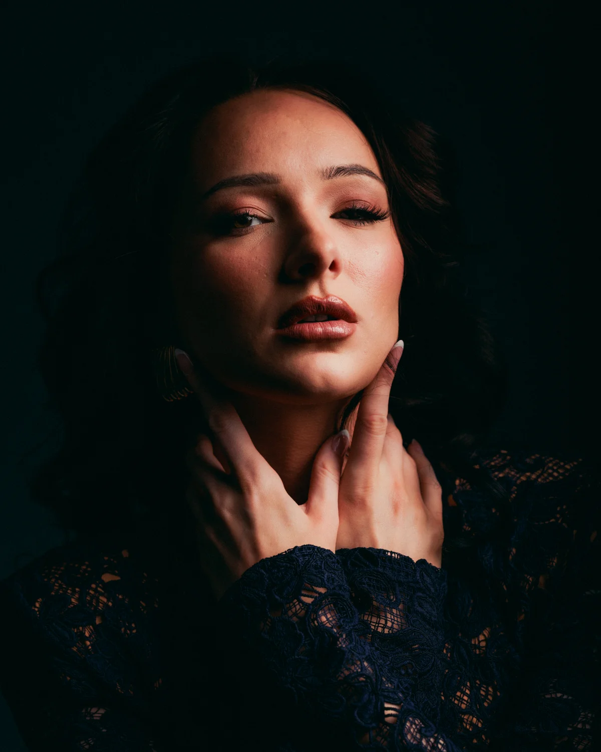 Moody close-up portrait of a woman with her hands on her neck, wearing dark lace against a black background.