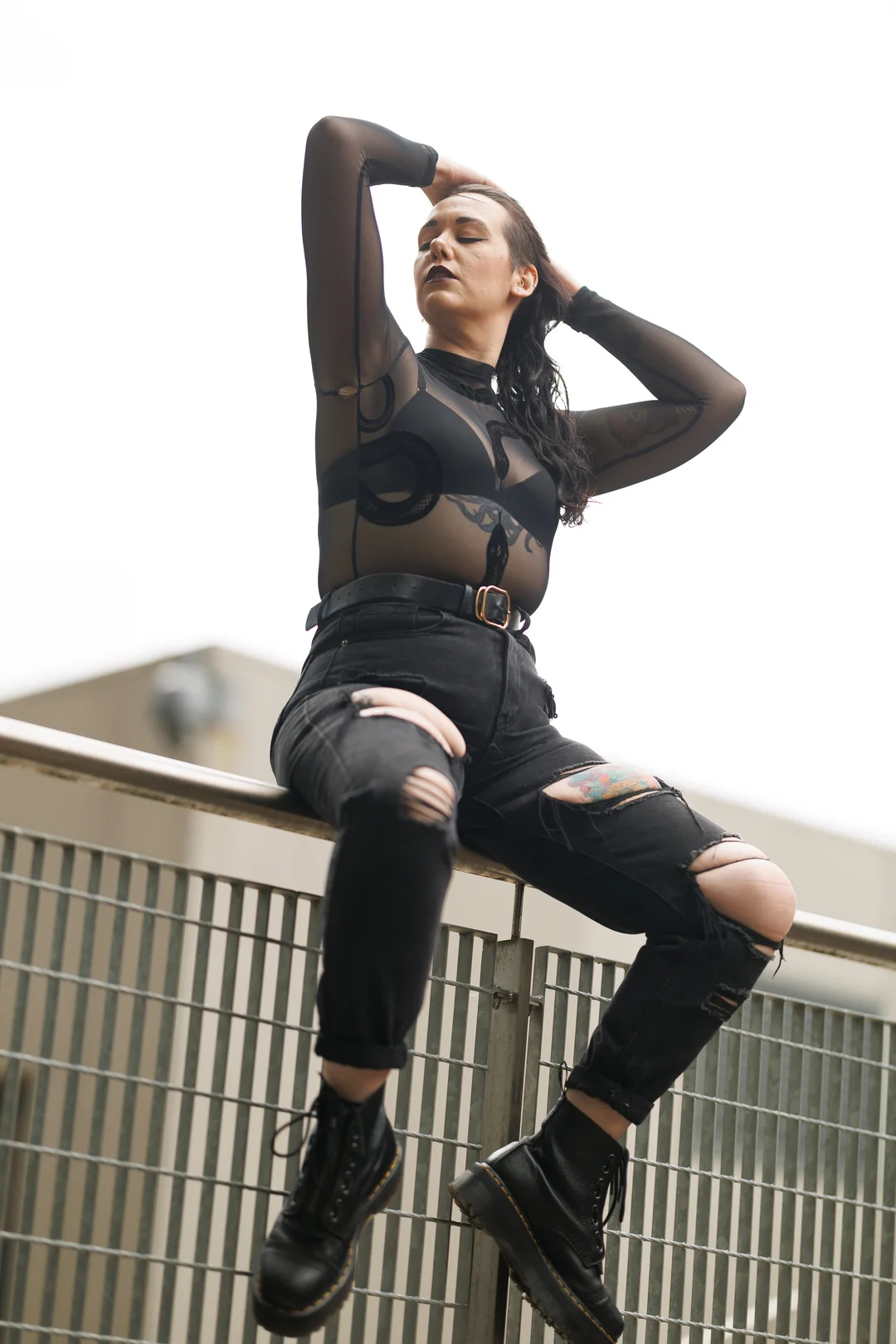 A woman in a black sheer top and ripped black jeans posing on a metal railing in an urban setting.