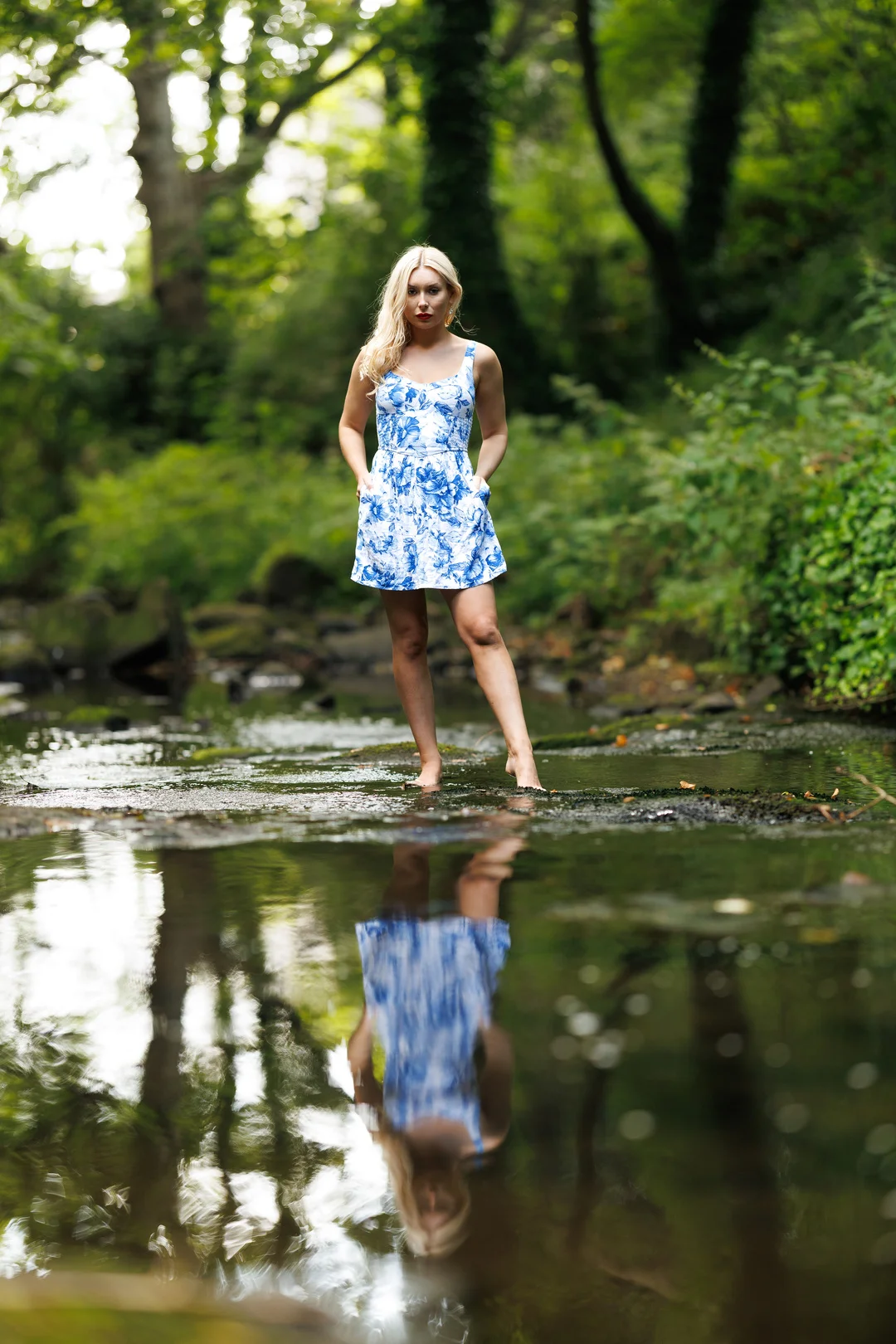 A woman in a blue and white floral dress standing in a shallow forest stream with a clear reflection in the water.