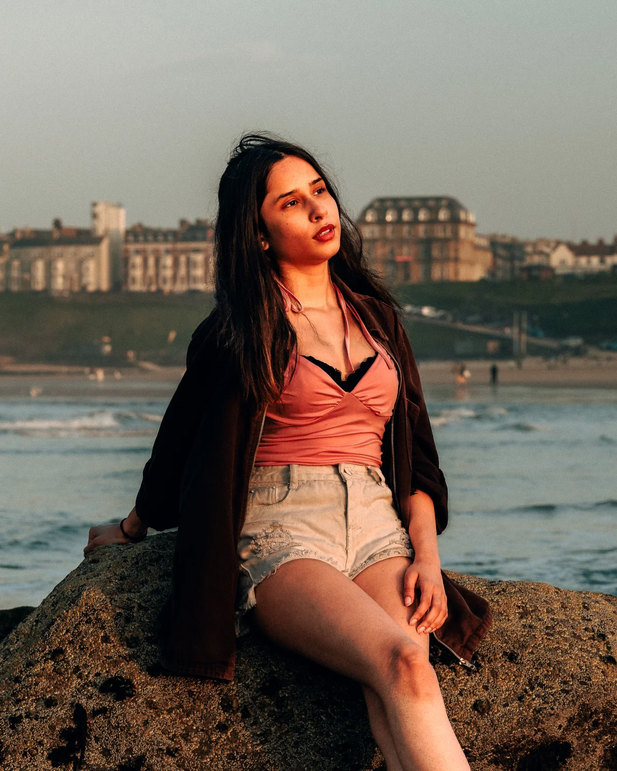A young woman with long dark hair sitting on a rock by the ocean at sunset, wearing a pink top and white shorts.