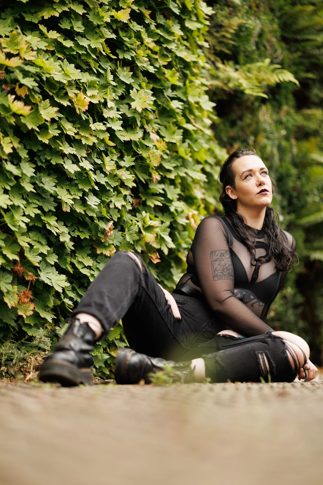 Person with dark hair and alternative style sitting on the ground against a lush green hedge.