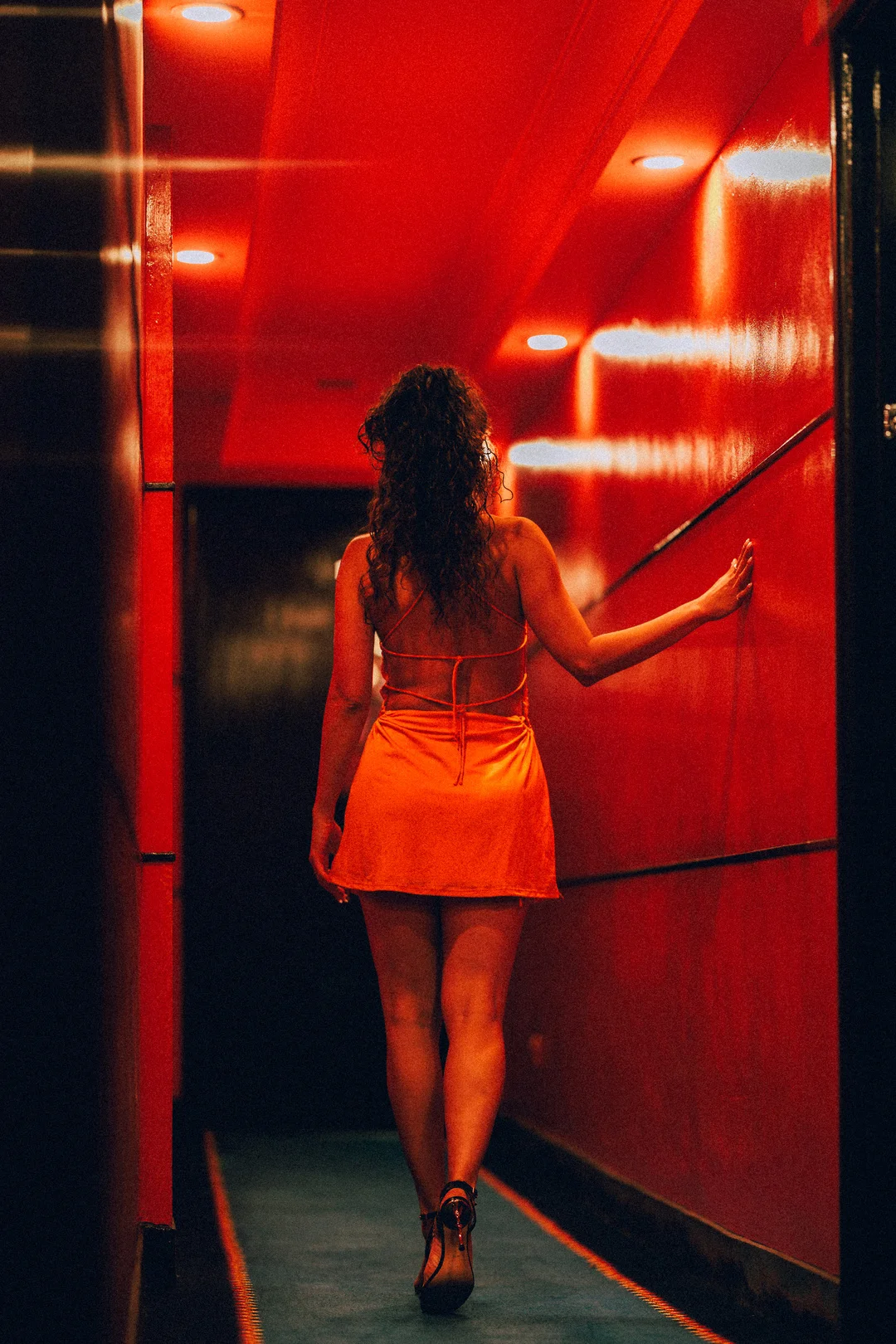 Woman in a short orange dress walking away through a moody, red-lit hallway.