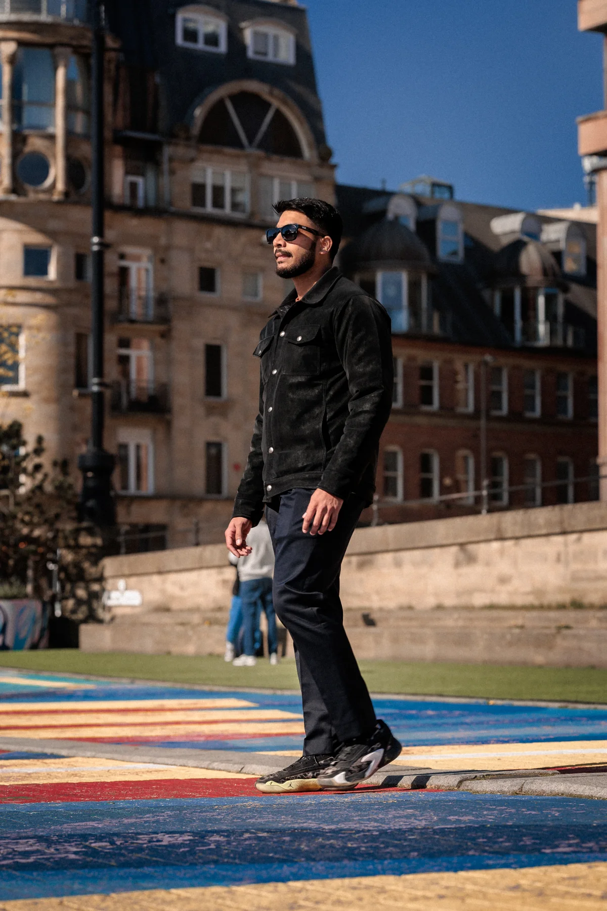 A man in a black jacket and sunglasses walking across a colorful rainbow crosswalk in an urban setting.