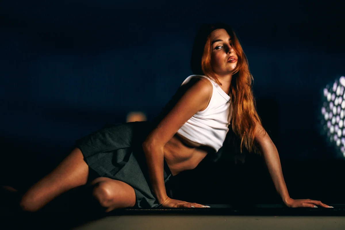 Woman with long red hair posing in a white tank top and dark skirt under dramatic, low-key lighting.