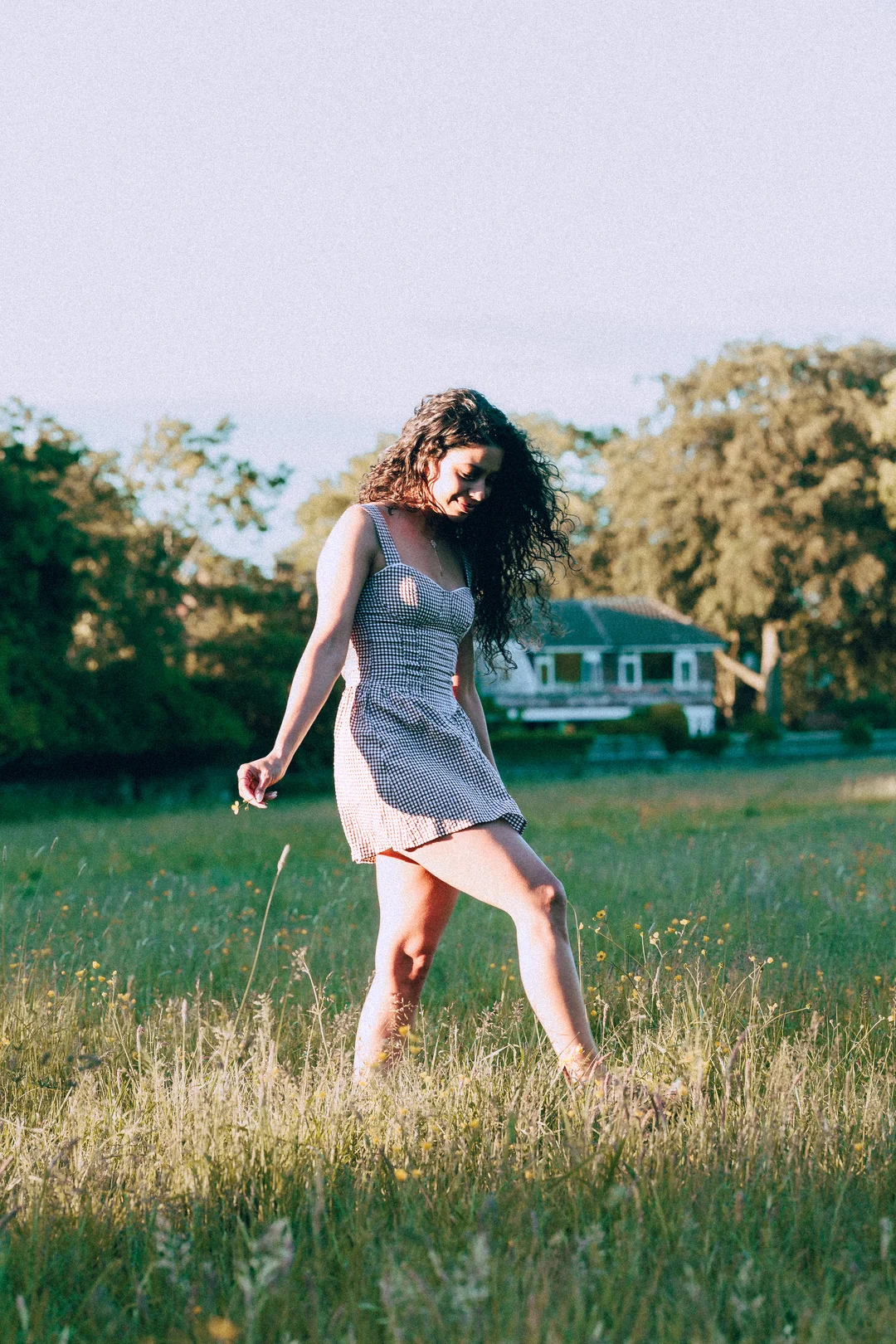 A woman with curly hair in a gingham dress walking through a sunny grassy field.