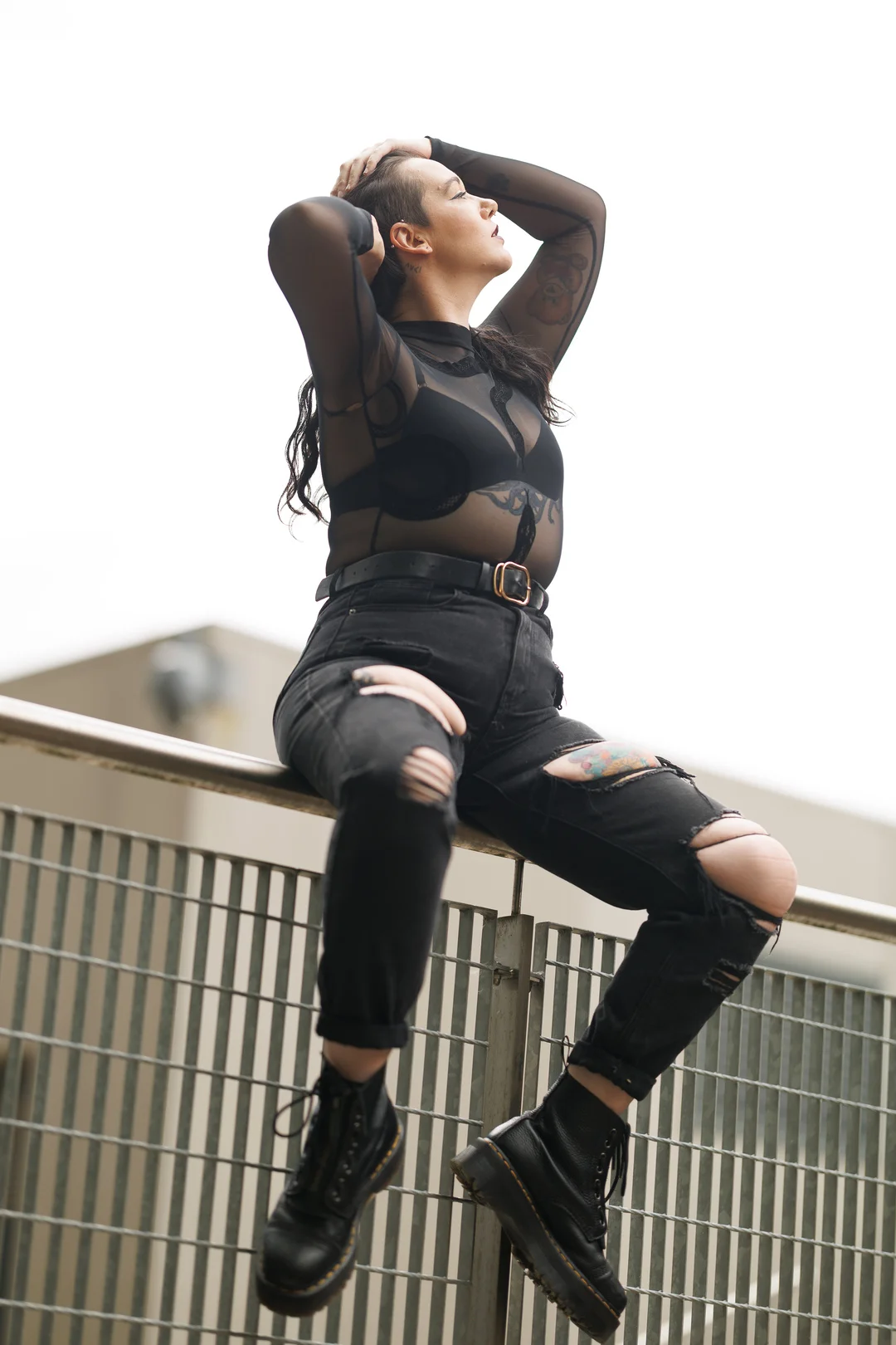 A woman in a sheer black top and ripped black jeans posing on a metal railing outdoors.