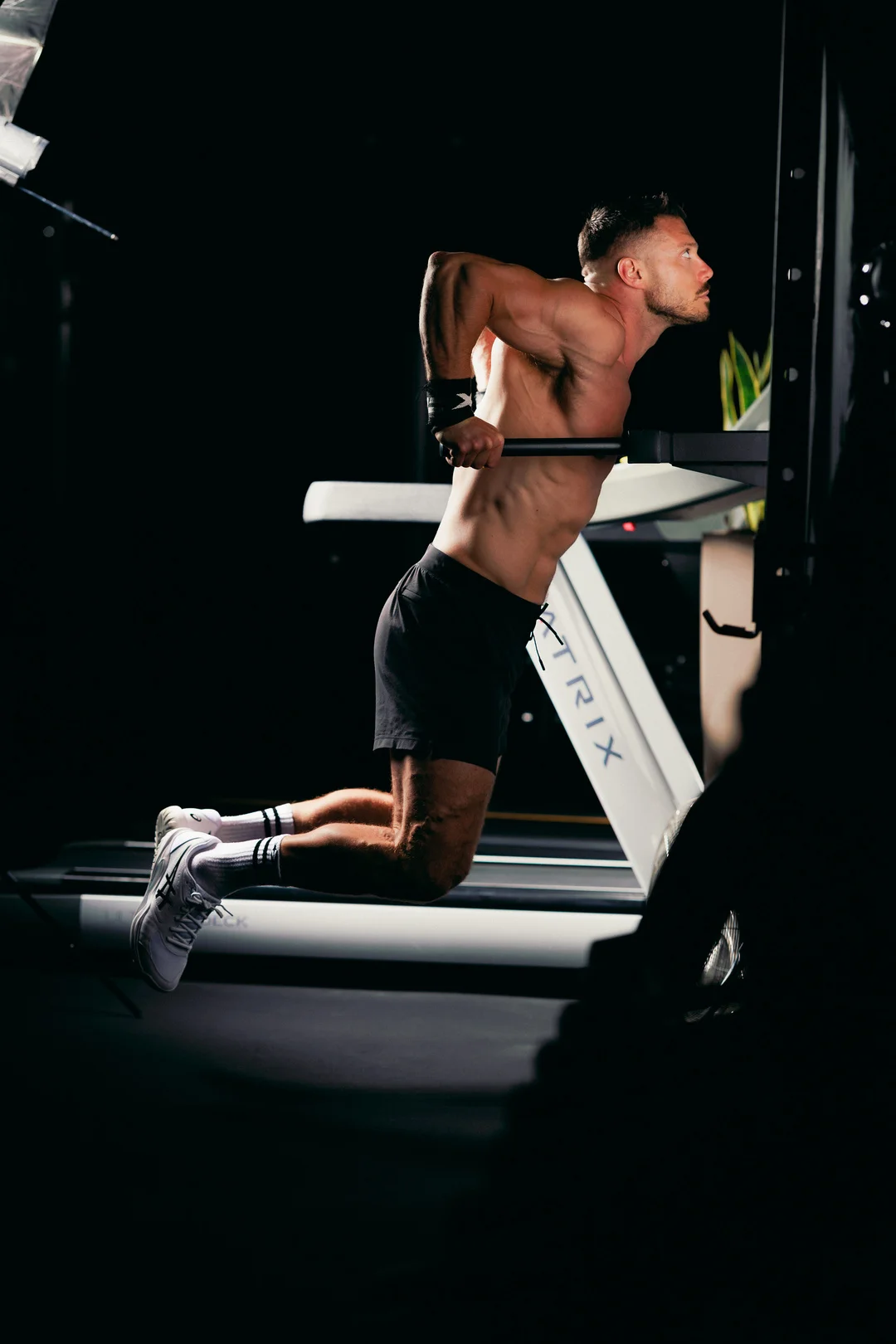 Shirtless man performing a dip on gym equipment with dramatic, high-contrast studio lighting.