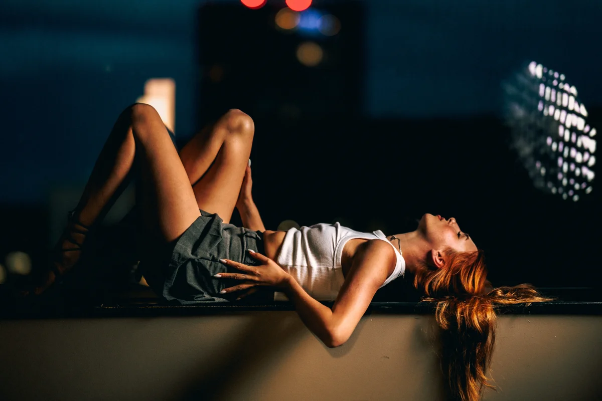 Woman with red hair lying on a ledge in a moody, nighttime urban setting, wearing a white top and grey skirt.