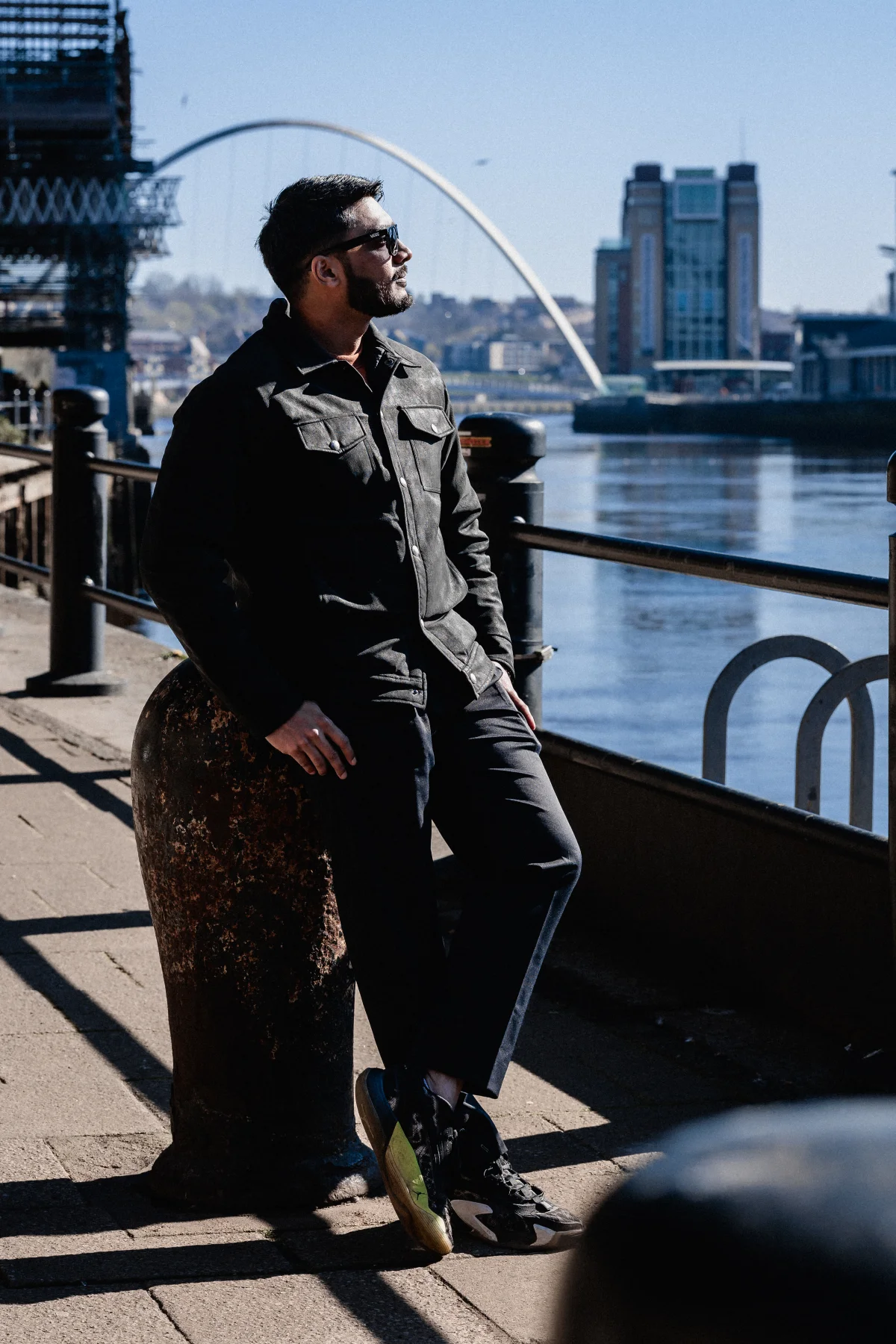 Man in a black jacket and pants posing by a river with a city skyline and bridge in the background.