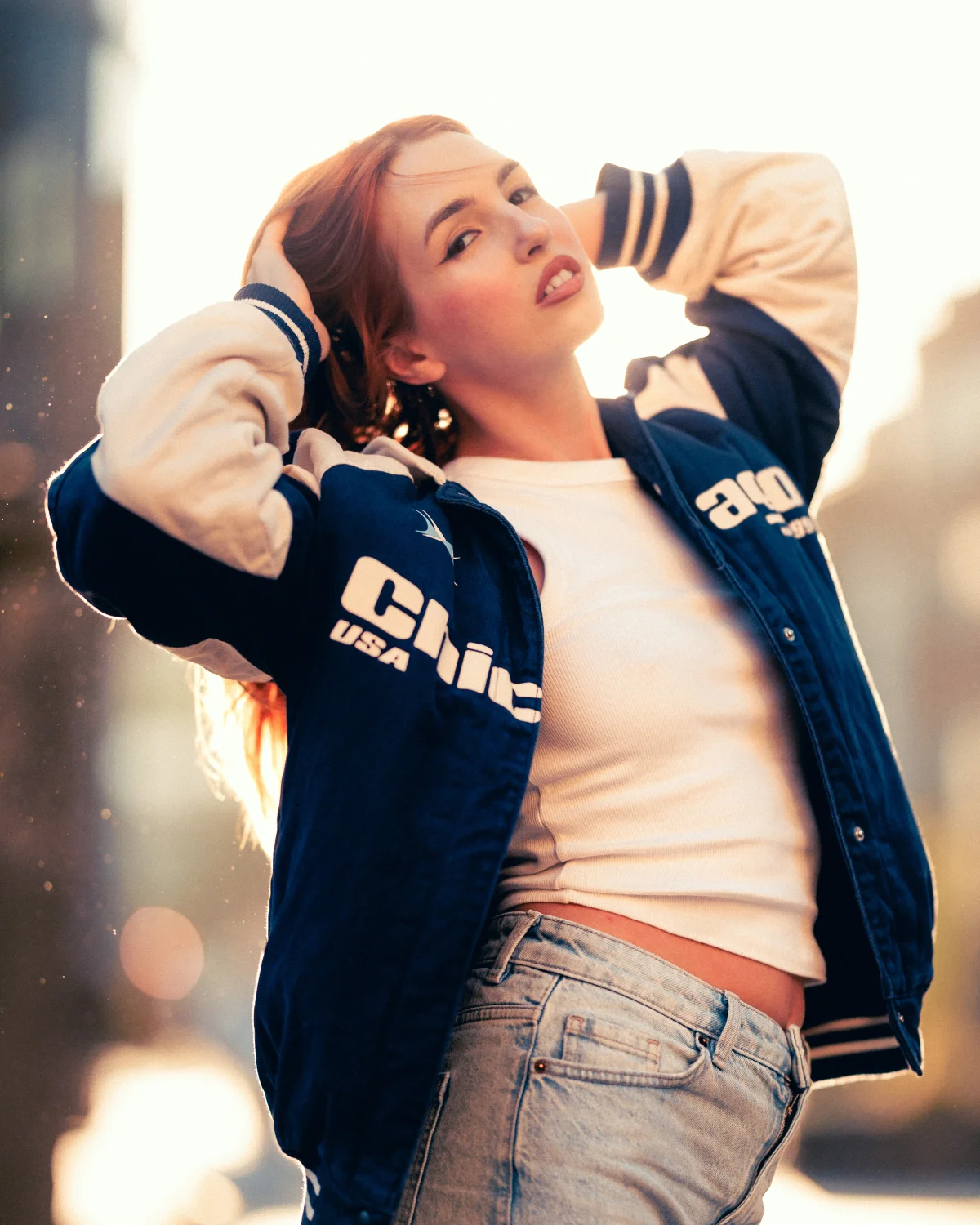 Young woman with red hair posing outdoors in a blue and white varsity jacket and light-wash jeans.