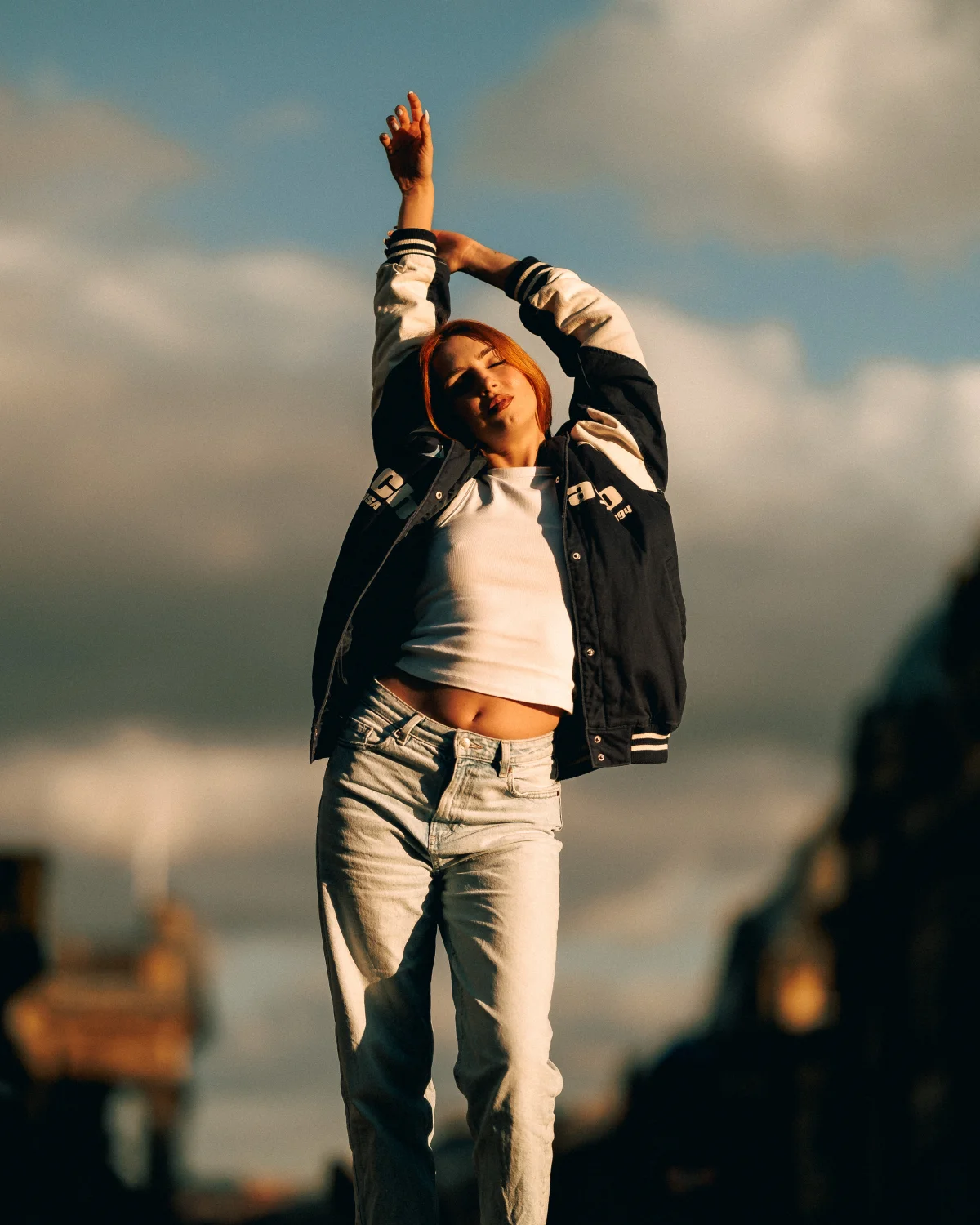 Woman with red hair posing with arms raised in a varsity jacket and jeans under a cloudy sky.