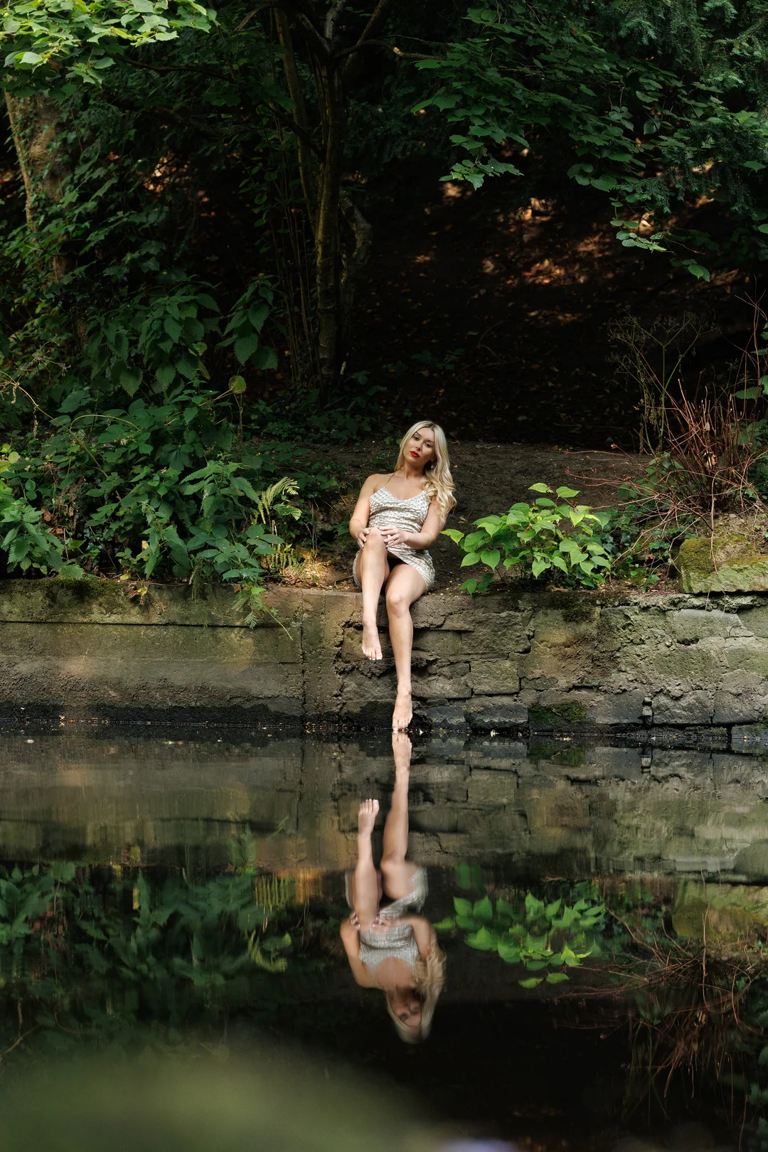 Blonde woman in a short dress sitting on a stone ledge by a pond with a clear reflection in the water.