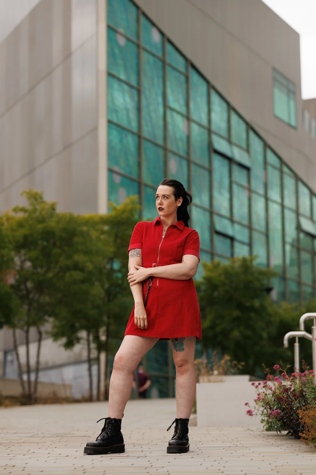 Woman in a short red zip-up dress and black boots posing in front of a modern glass building.