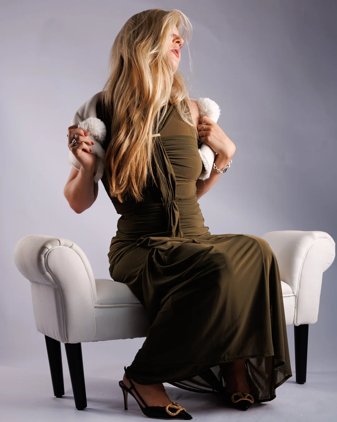 Woman with long blonde hair in an olive green gown and white fur wrap posing on a white chaise lounge.