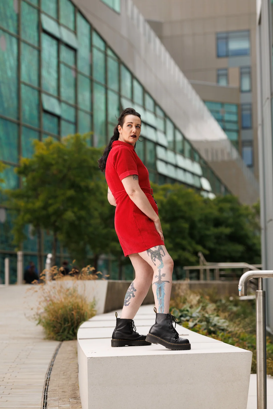 Woman with tattoos in a red dress and black boots posing before a modern glass building.