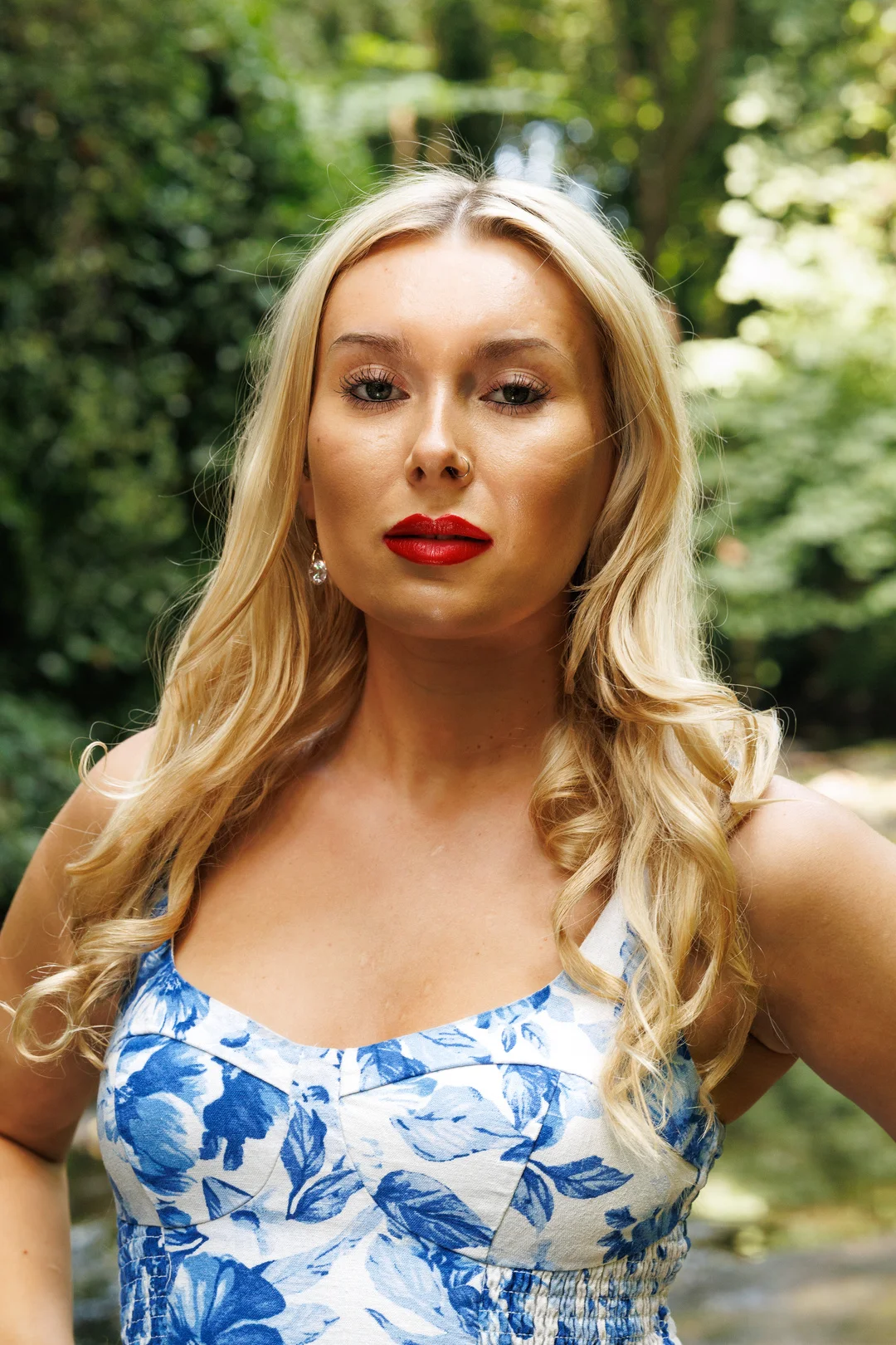 Blonde woman with red lipstick in a blue floral dress posing against a blurred green forest background.