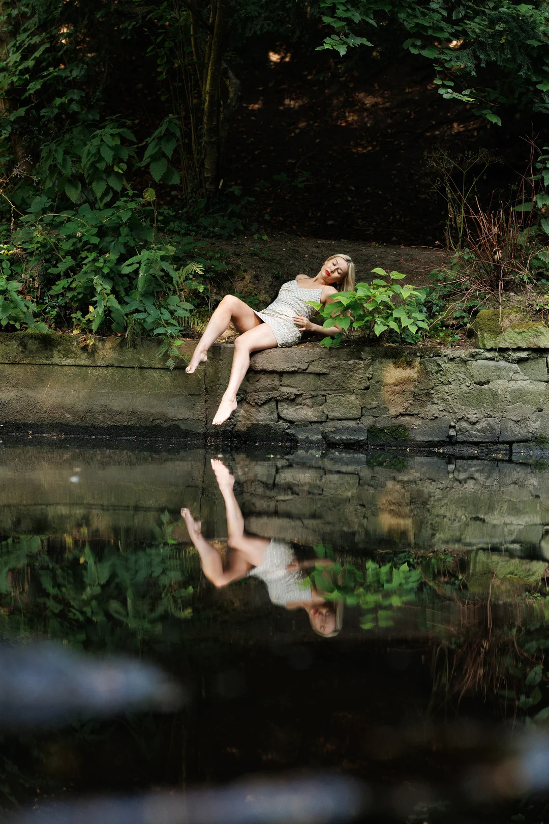 Woman in a short silver sequin dress reclining on a stone wall by a still pond, reflecting her image.