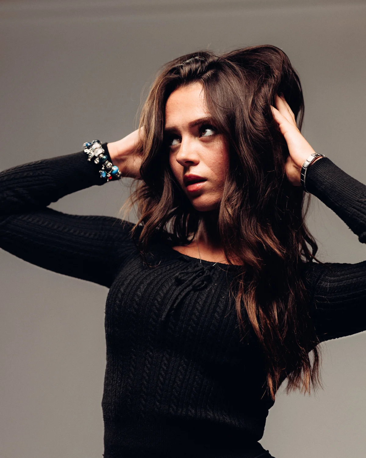 A woman with long brown hair posing with her hands in her hair, wearing a black ribbed sweater against a grey background.