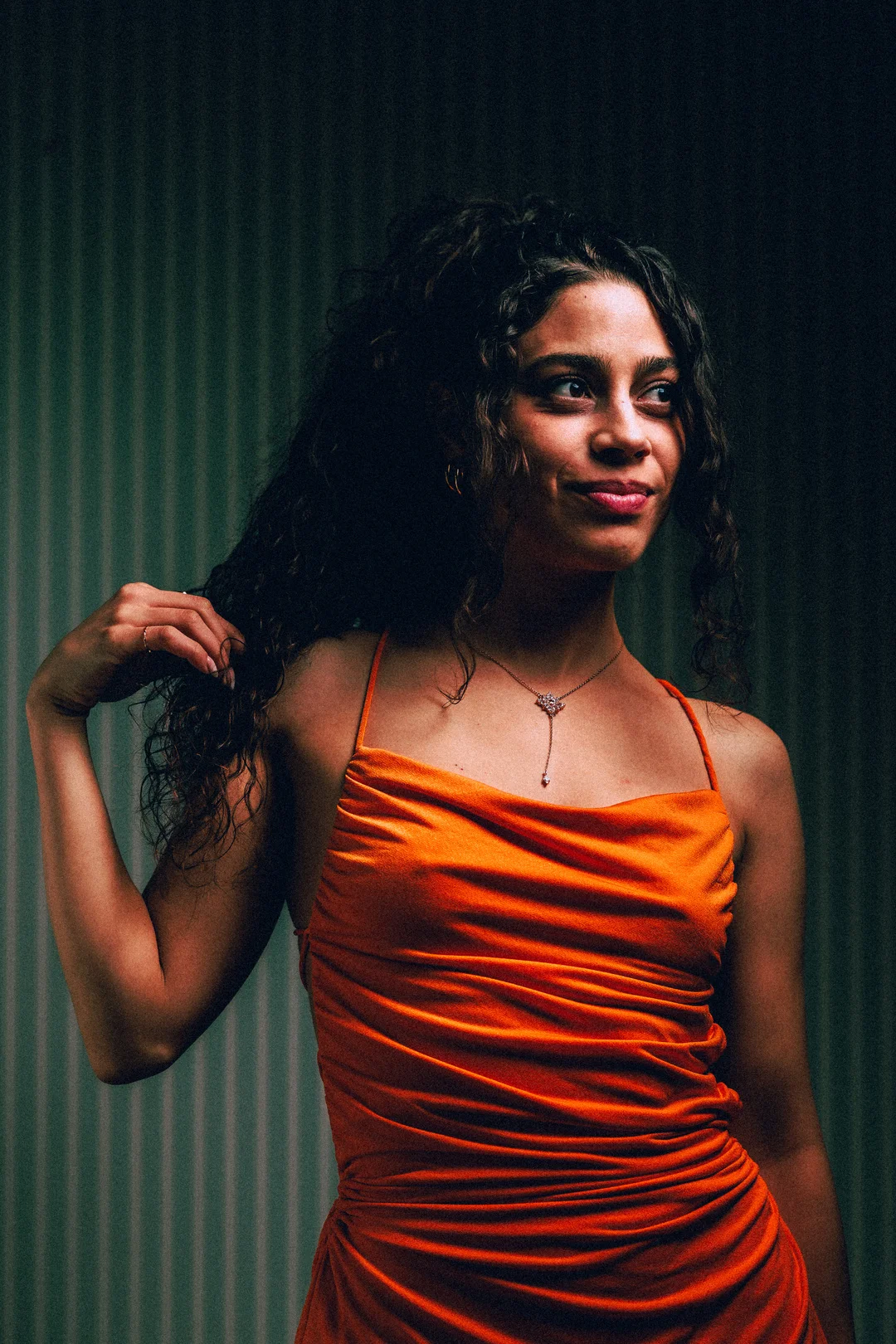 A woman with curly dark hair wearing an orange satin dress, looking off-camera against a dark ribbed background.