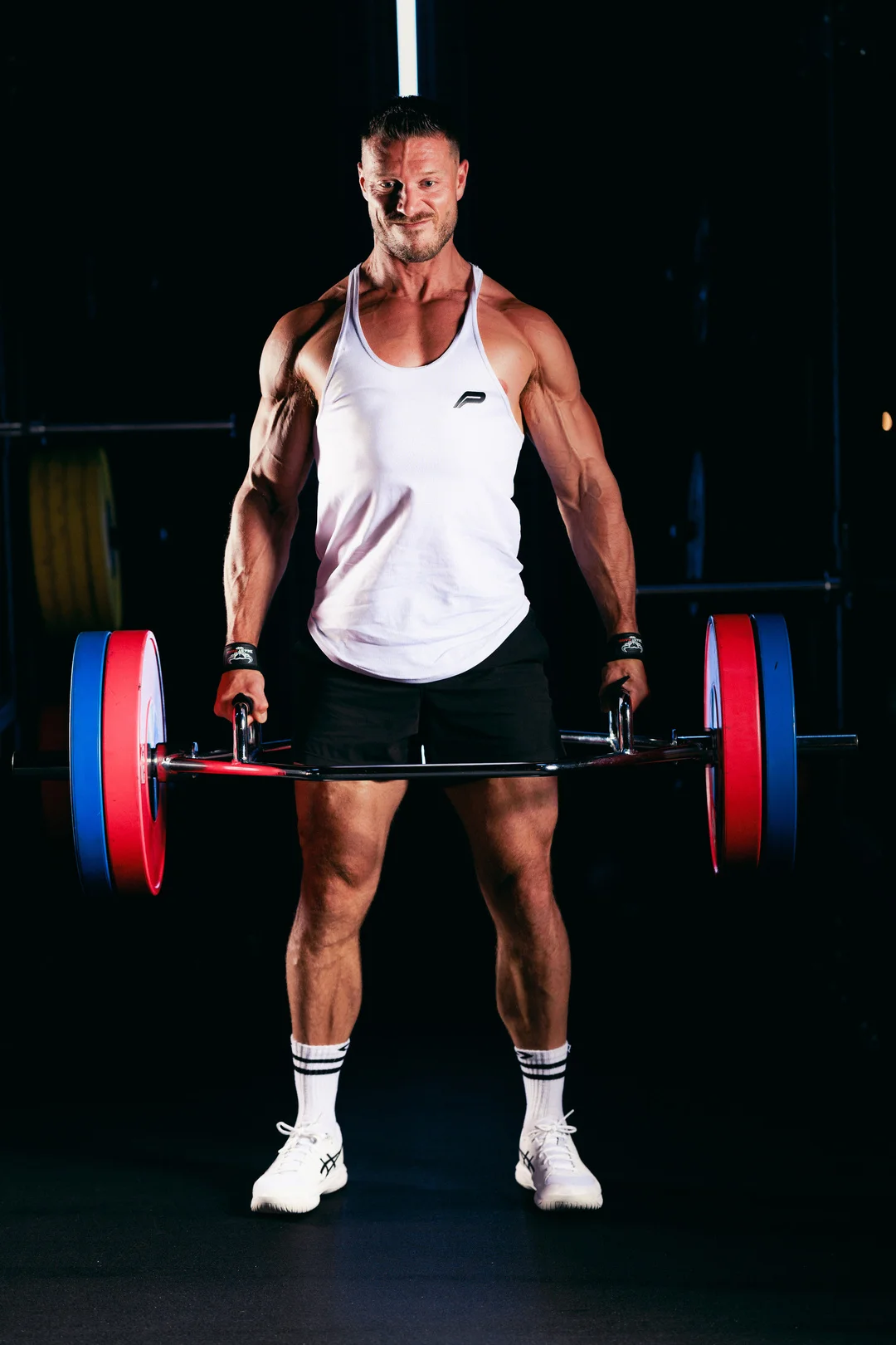 Muscular man in a white tank top posing with a barbell in a dark gym setting.