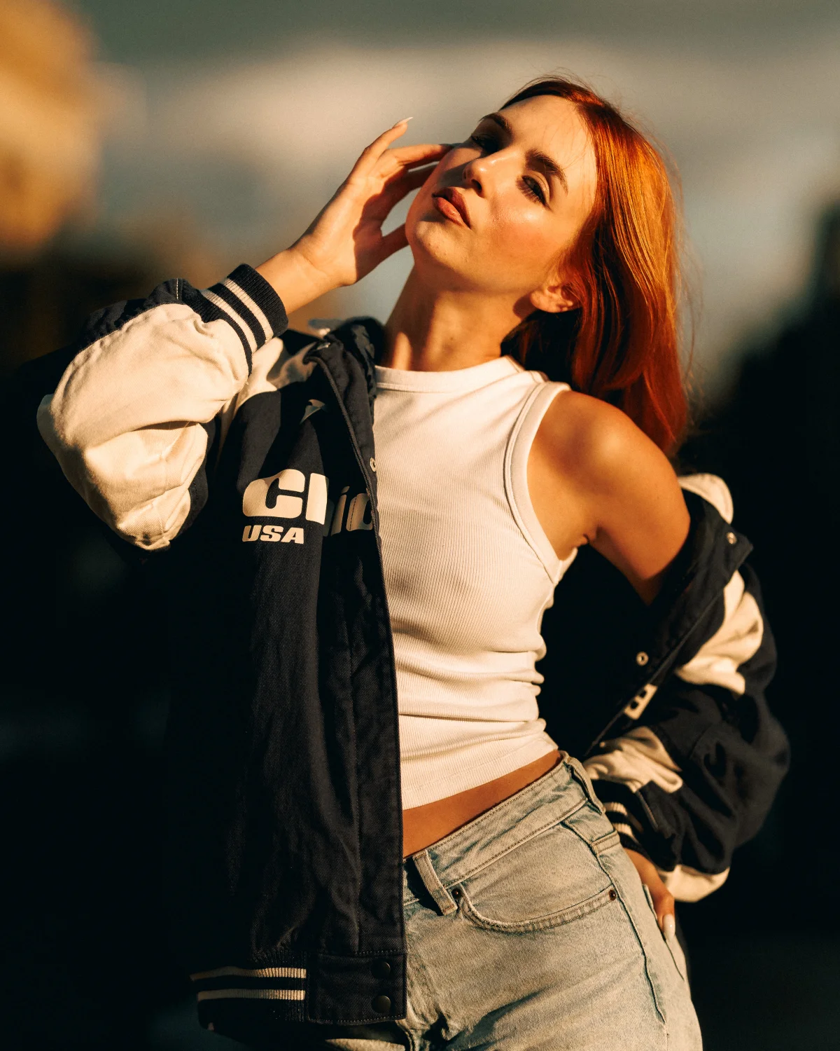 Woman with red hair posing in a varsity jacket and white crop top during golden hour.