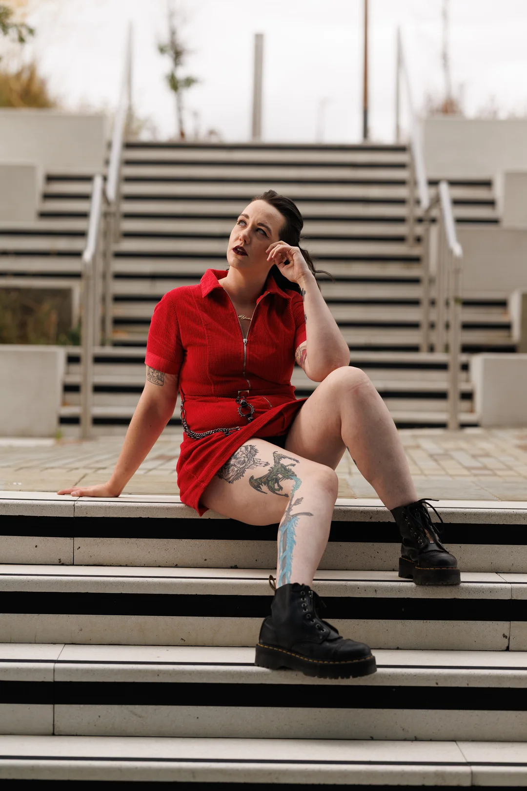 A person in a red zip-up dress and black boots sitting on outdoor stairs, looking away.