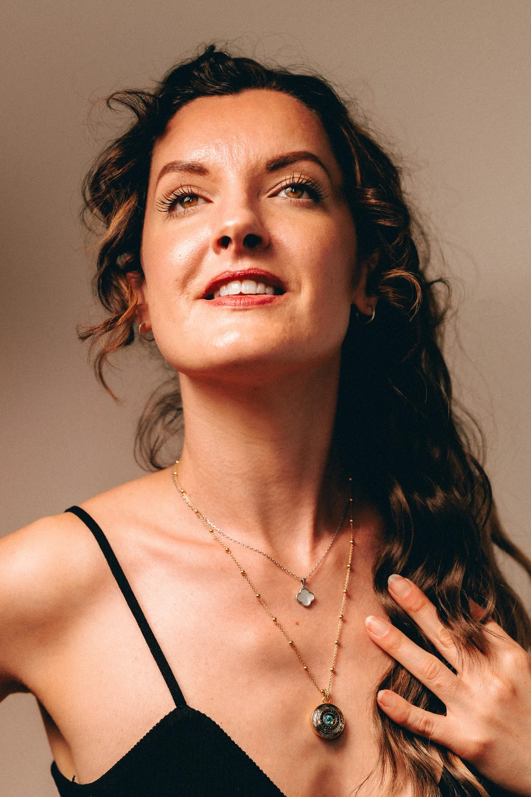 Close-up portrait of a woman with curly hair looking up, wearing gold necklaces against a neutral backdrop.