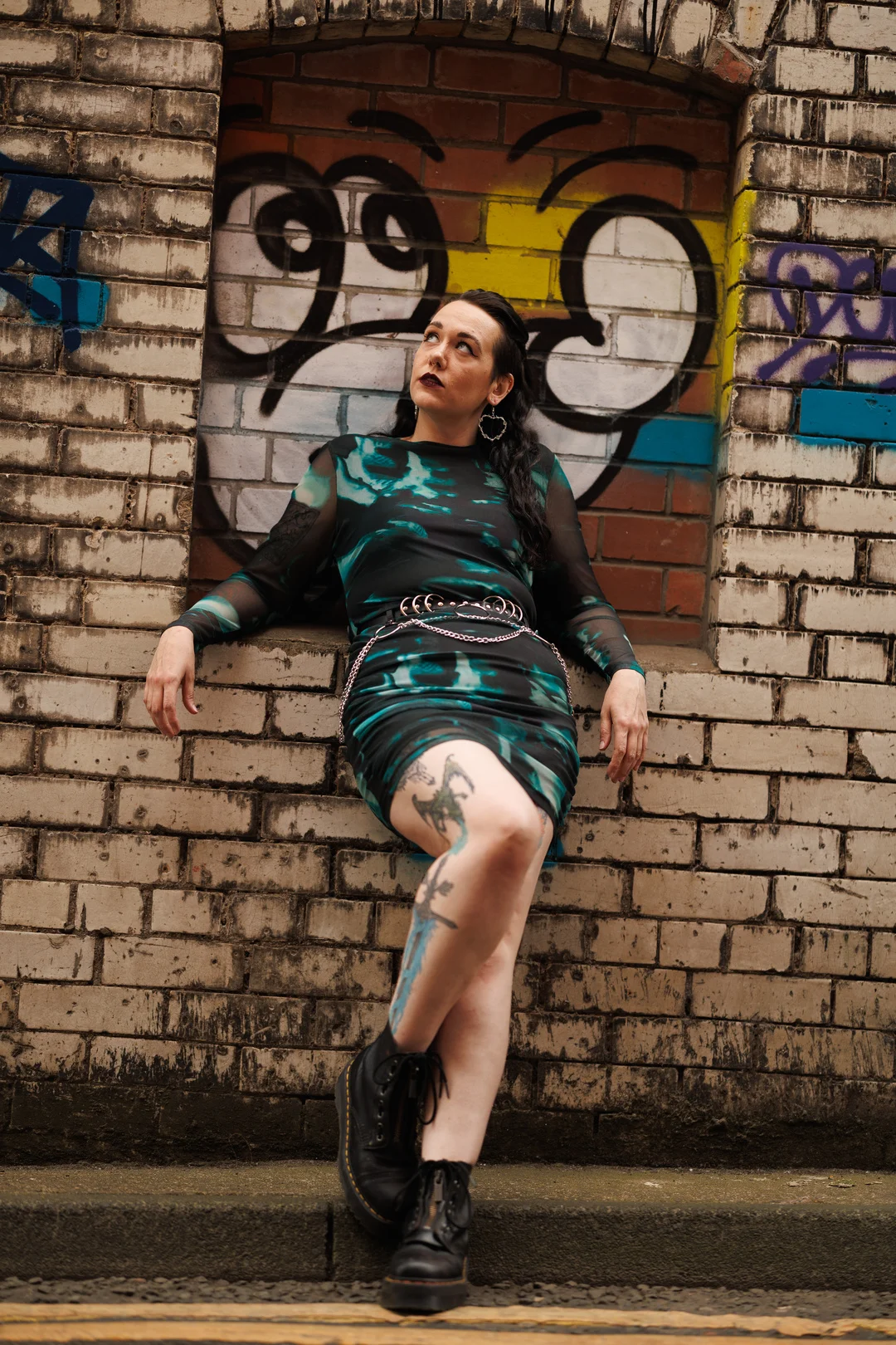 A woman with tattoos and dark hair posing in a teal dress against a graffiti brick wall.