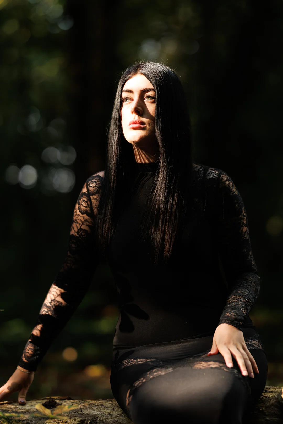 Moody outdoor portrait of a woman with long black hair wearing a black lace outfit in a shaded forest.