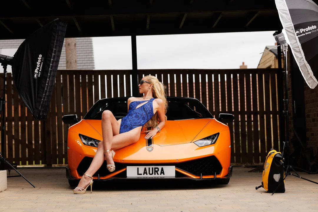 Blonde woman in a blue swimsuit posing on an orange Lamborghini with studio lighting.