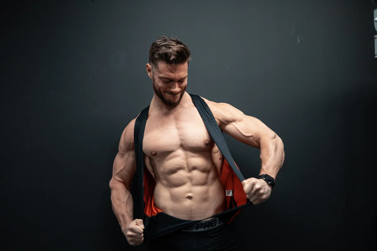 Muscular man in a black tank top pulling it up to reveal his abs, smiling against a dark studio background.