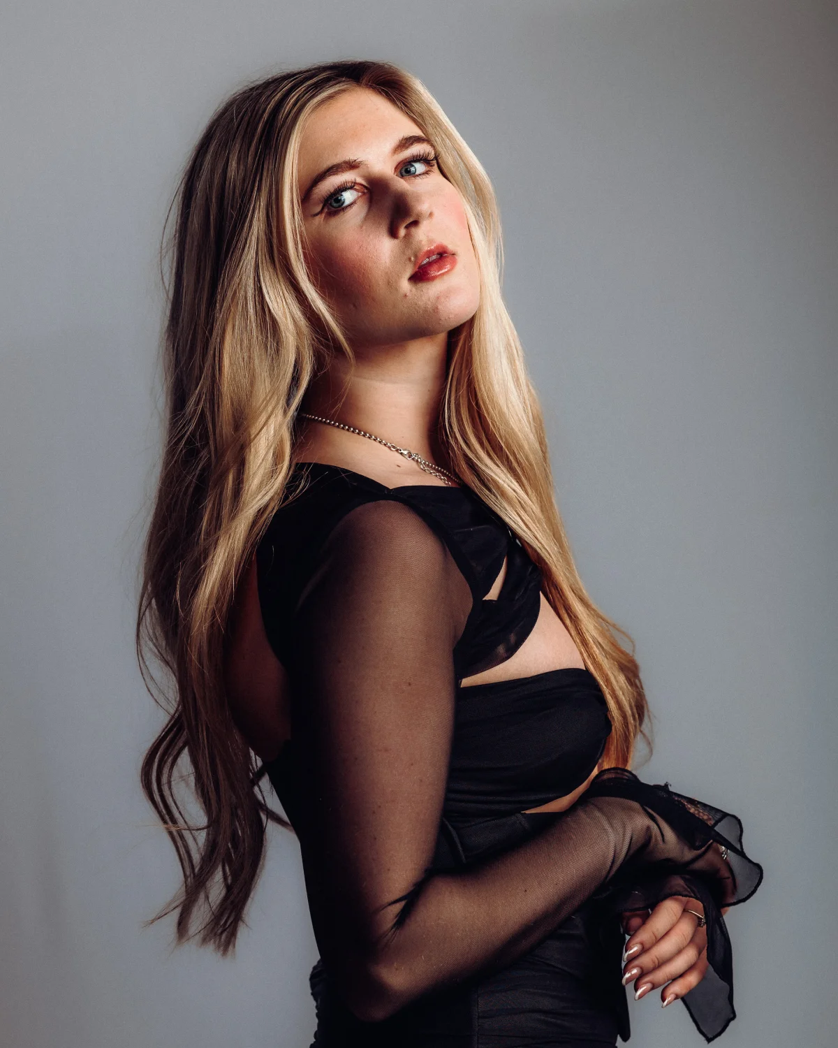 A young woman with long blonde hair in a black sheer outfit posing against a grey studio background.