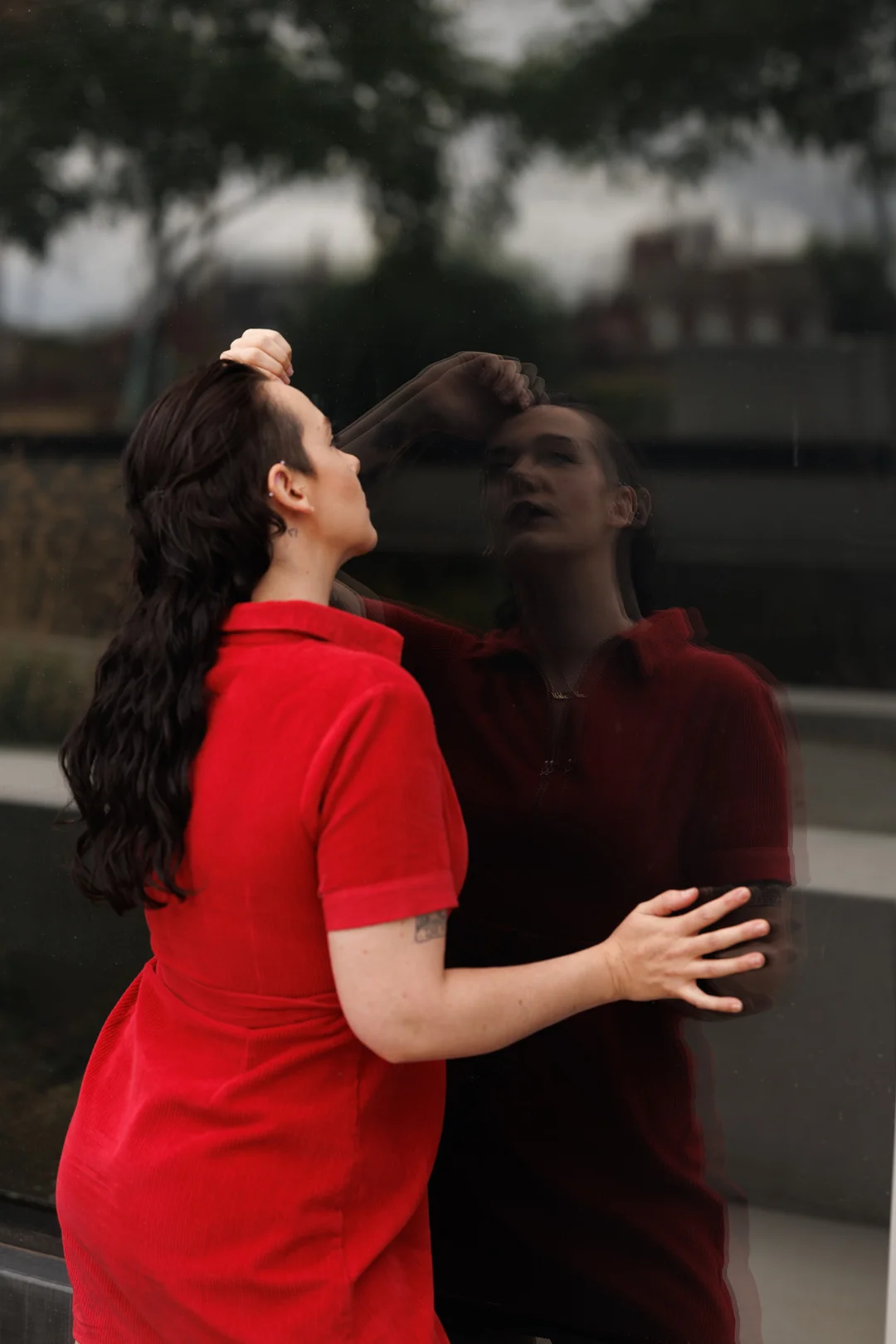 A woman in a red dress touching her reflection in a dark glass window outdoors.
