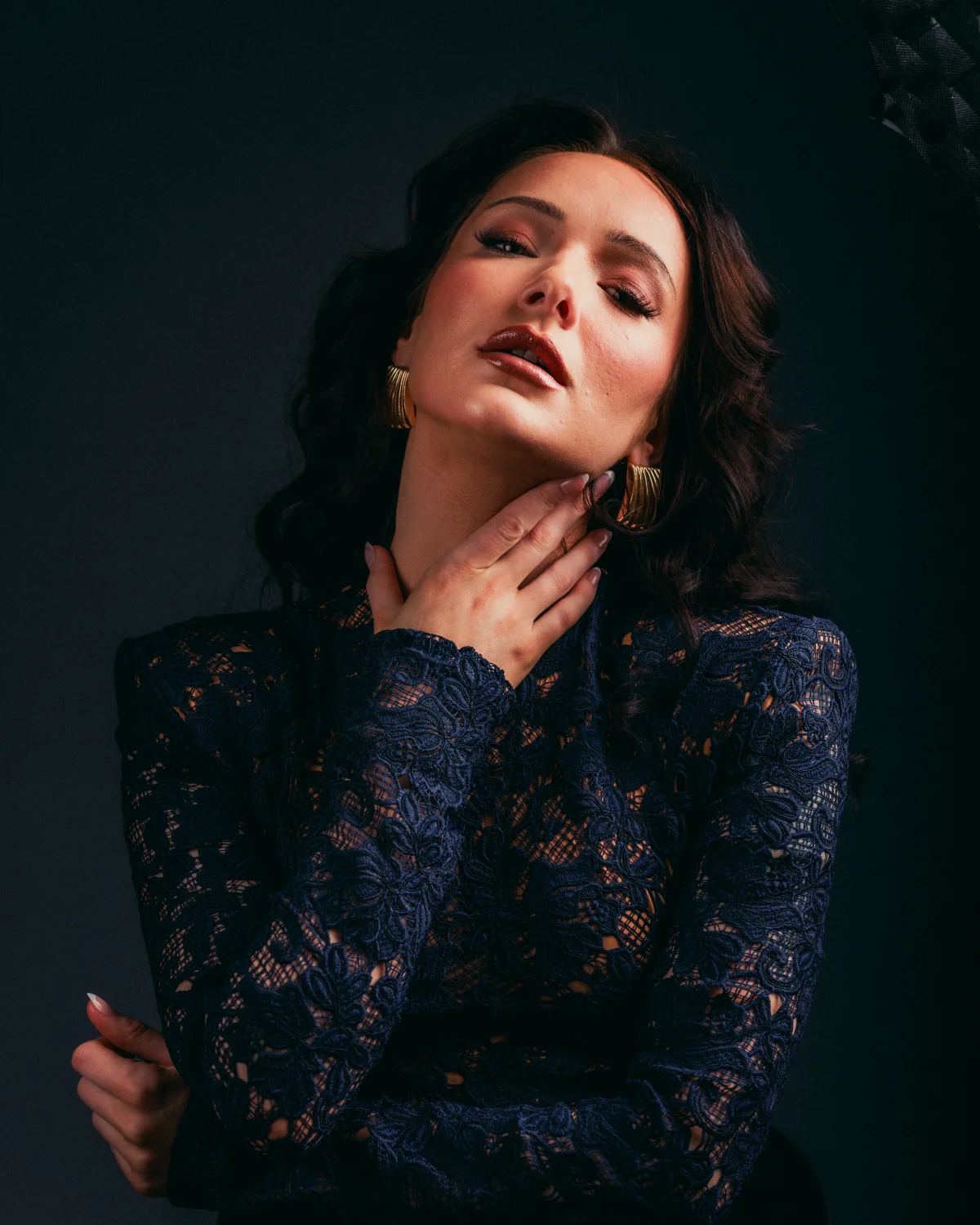 A woman with dark hair and gold earrings in a navy blue lace top, posing against a dark background.