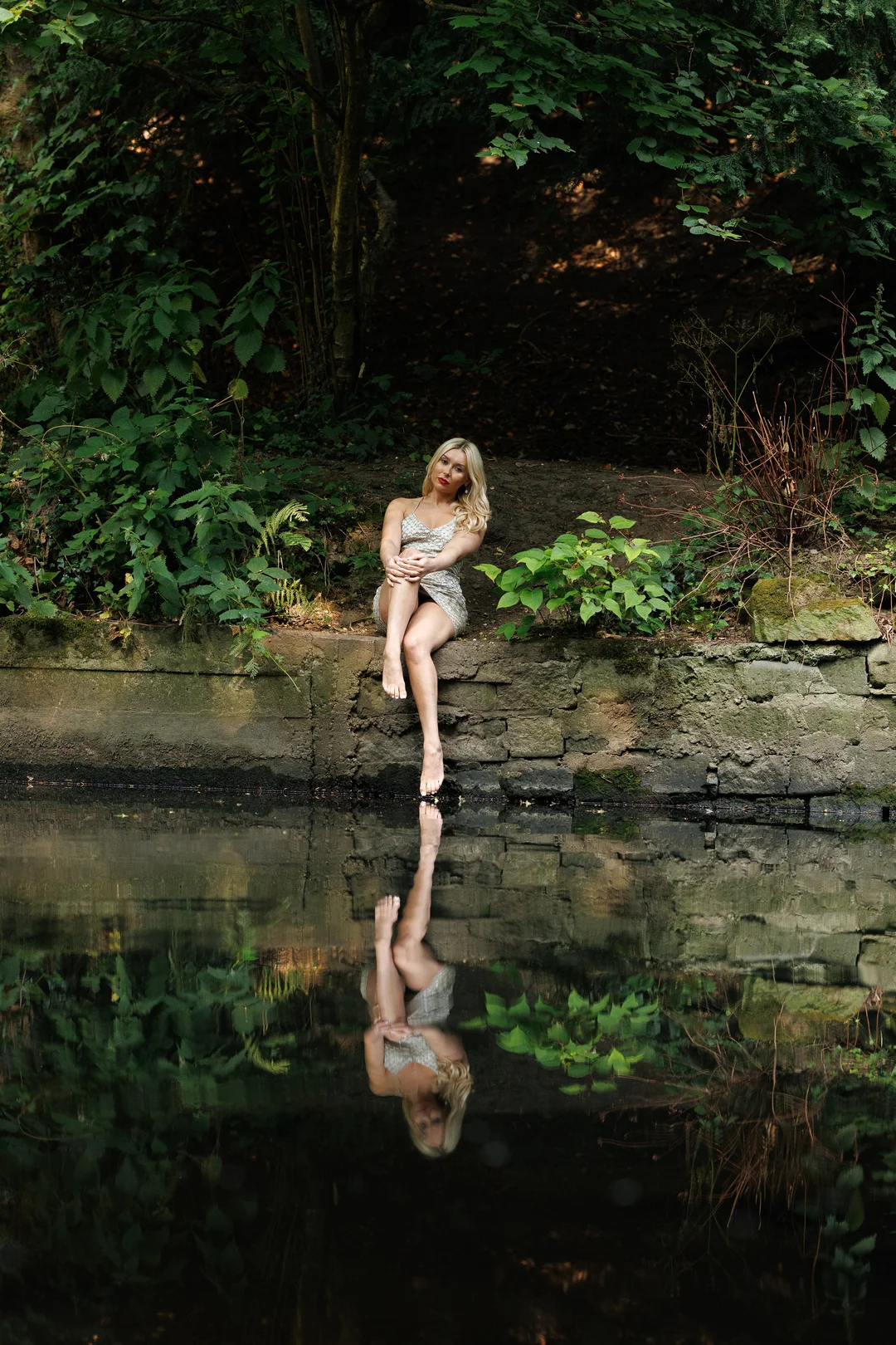 Blonde woman in a short sparkly dress sitting on a stone ledge by a reflecting pond in a forest.