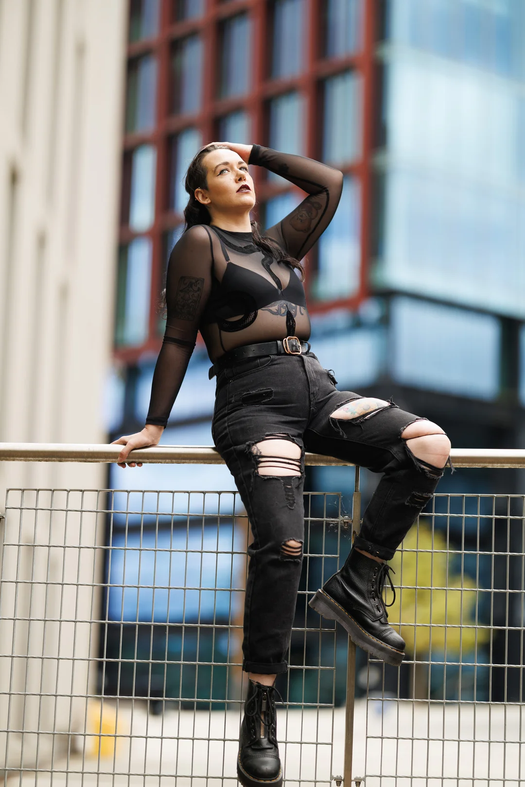 A person in a sheer black top and ripped black jeans posing against a railing in an urban setting.