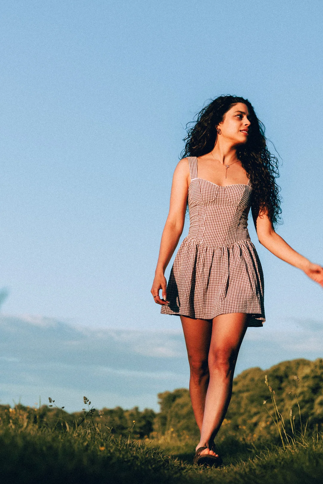A woman with long curly hair in a red and white gingham mini dress standing in a grassy field under a blue sky.