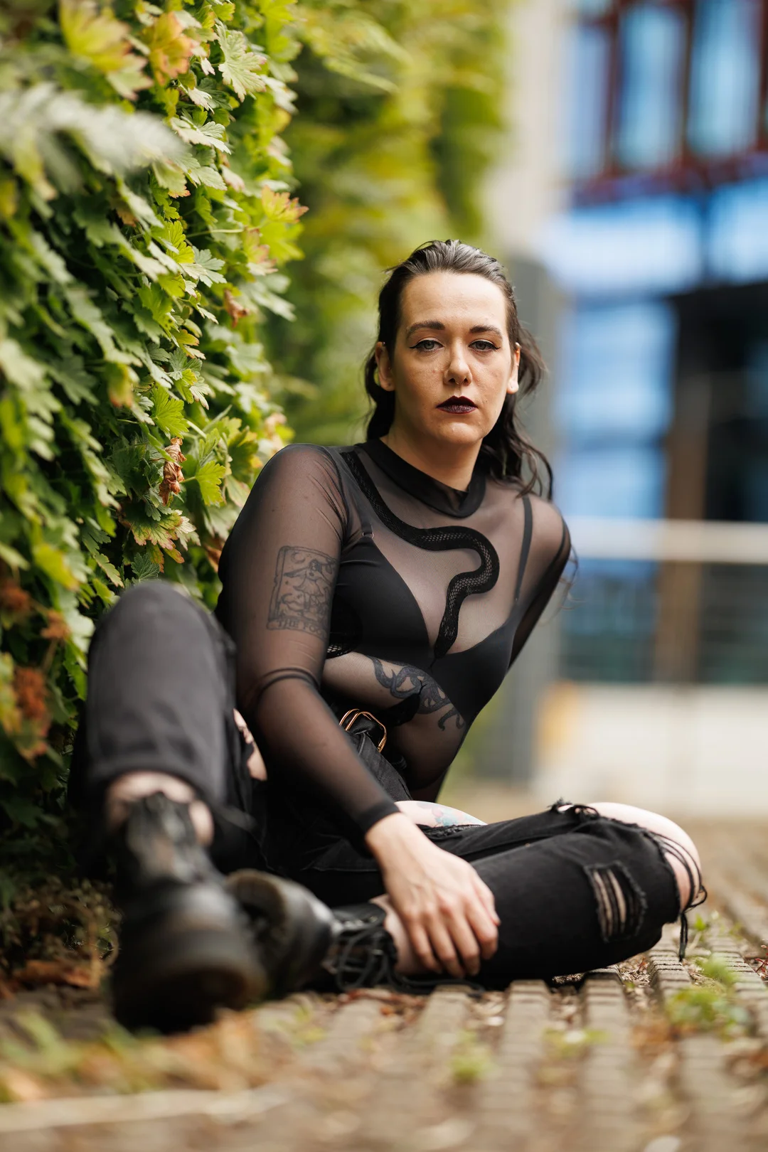 A person with dark hair and makeup, wearing a black sheer top and ripped jeans, sitting outdoors against a green hedge.