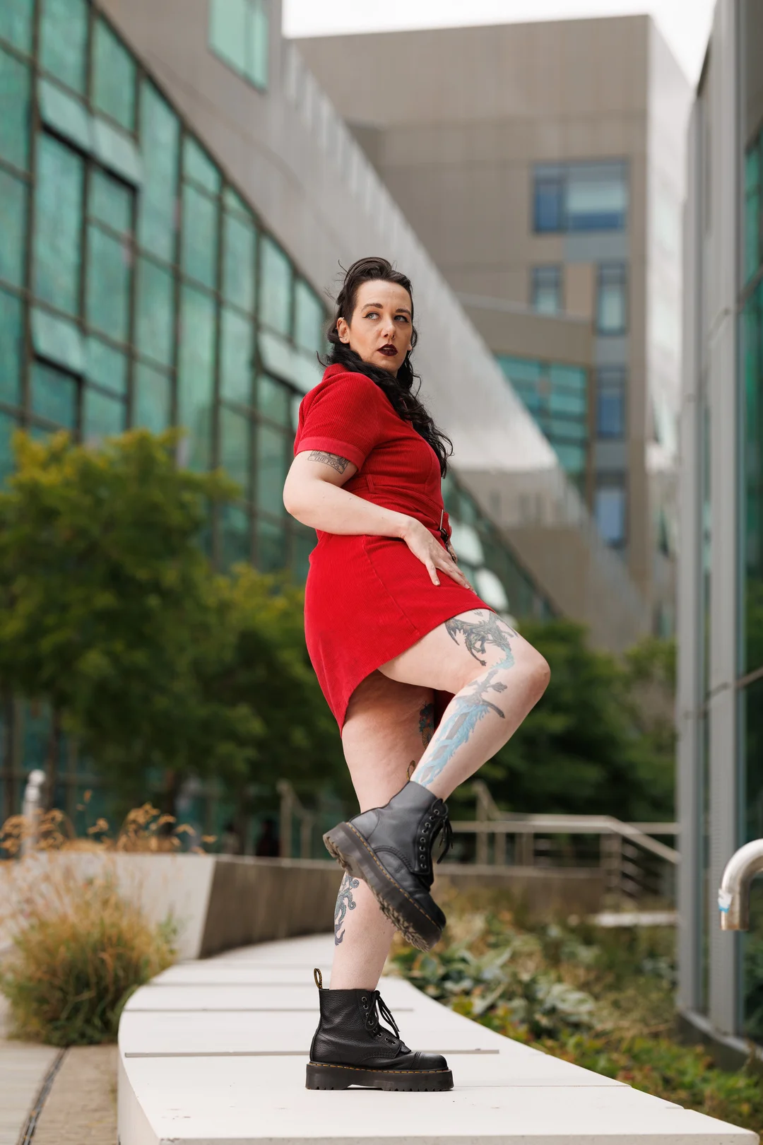 Woman with tattoos in a red dress and black boots posing in a modern urban setting.